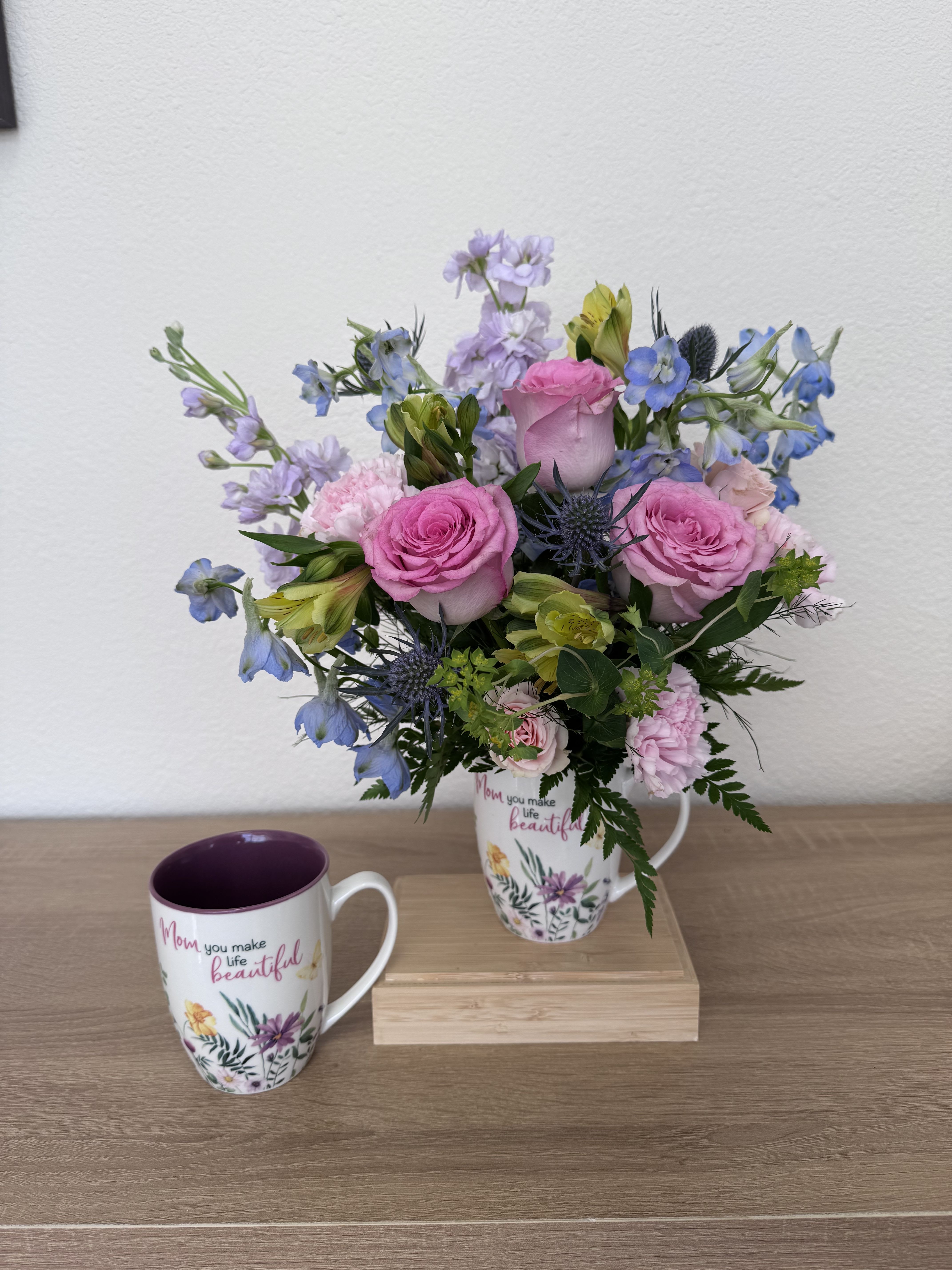 Mom's Garden Mug - Soft, garden-fresh pink roses and pale pink carnations mingle with airy blue delphinium-like blooms and lavender accents in a white ceramic mug-an ideal Mother's Day or birthday flower arrangement that reads feminine and affectionate. The mug's floral graphics and the heartfelt message, "Mom you make life beautiful," make this bouquet an instant gift that says she's seen and loved; the light, springlike palette gives a cheerful, handmade feel that delights at first glance.  Gathered in a white ceramic mug and shown with a matching mug beside it, the composition is loose and airy with a natural, just-picked texture from the mixed roses, alstroemeria, carnations, and soft filler blooms. The arrangement sits naturally on a wooden riser for display; colors include pink, blue, purple, and white for a soothing, complementary mix that suits a kitchen counter, bedside table, or office desk.  Perfect for the mom who likes fresh, feminine blooms-whether for Mother's Day, a birthday, or "just because"-this keepsake mug doubles as a small everyday reminder of your affection. Arrange for local delivery or pickup; to keep blooms vibrant, change water every other day and trim stems slightly when refreshing the vase.
