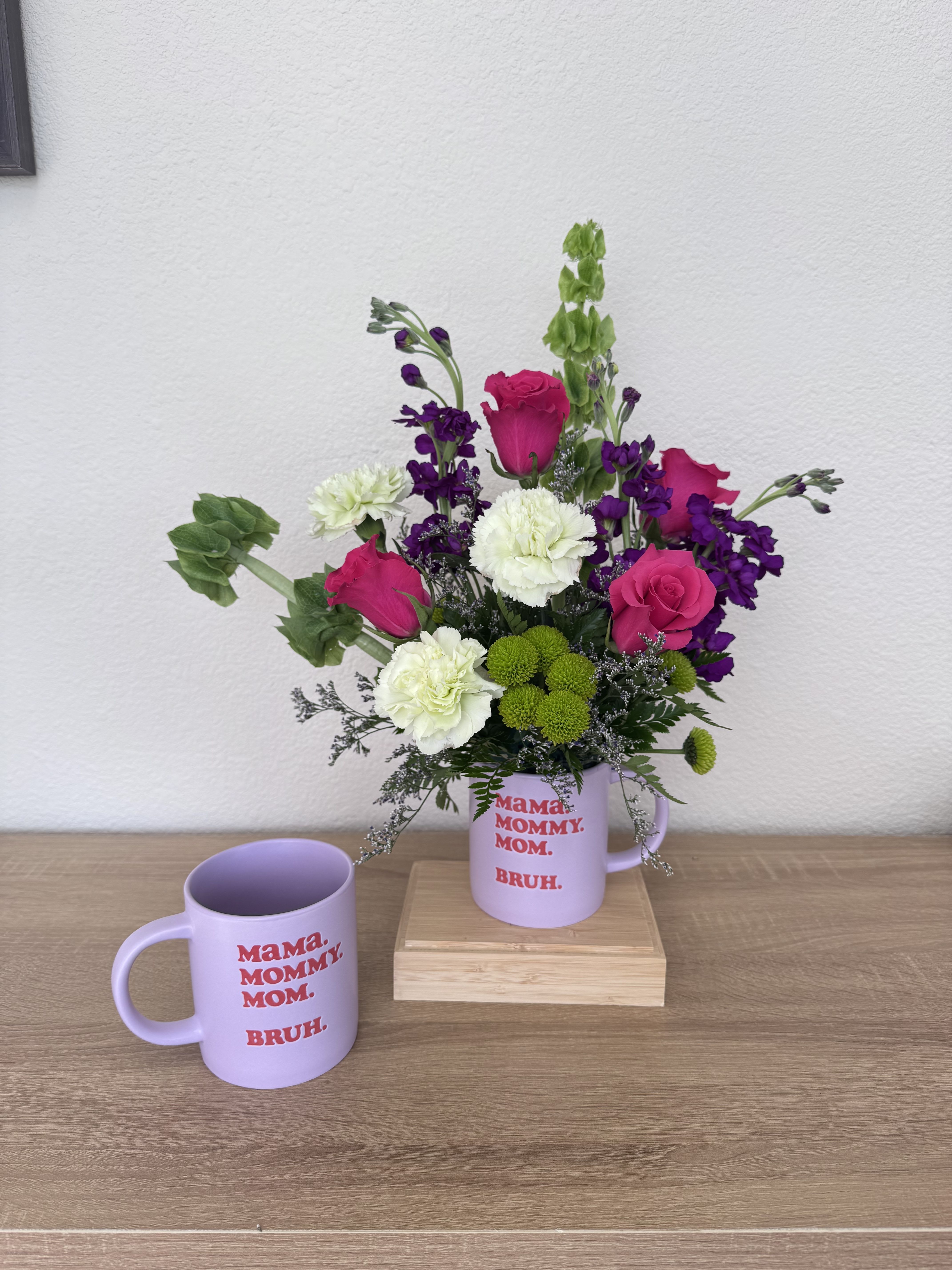 Bright Love for Mom - Hot pink roses and airy purple stock make a bright, joyful statement in this flower arrangement for Mother's Day or a mom's birthday. The upright, full design is layered with white carnations and little green chrysanthemum blooms that create playful contrast and a lively texture-perfect for the person you want to celebrate and make feel seen.  Arranged in a lavender ceramic mug with bold red text and placed on a wooden riser, the composition feels cheerful and informal-like a warm kitchen hello or a sunny bedside surprise. The purple stock adds vertical movement while the roses and carnations fill the eye; a few trailing accents bring a relaxed, hand-tied charm. The mug is reusable, making this both a bouquet and a keepsake.  Give this to a mom who delights in bright color and casual, homey style; it's the kind of gift that reads "thank you" and "I love you" at once. Order with confidence for delivery on Mother's Day or a birthday-no special care beyond fresh water in the mug will keep the blooms looking their best for several days.