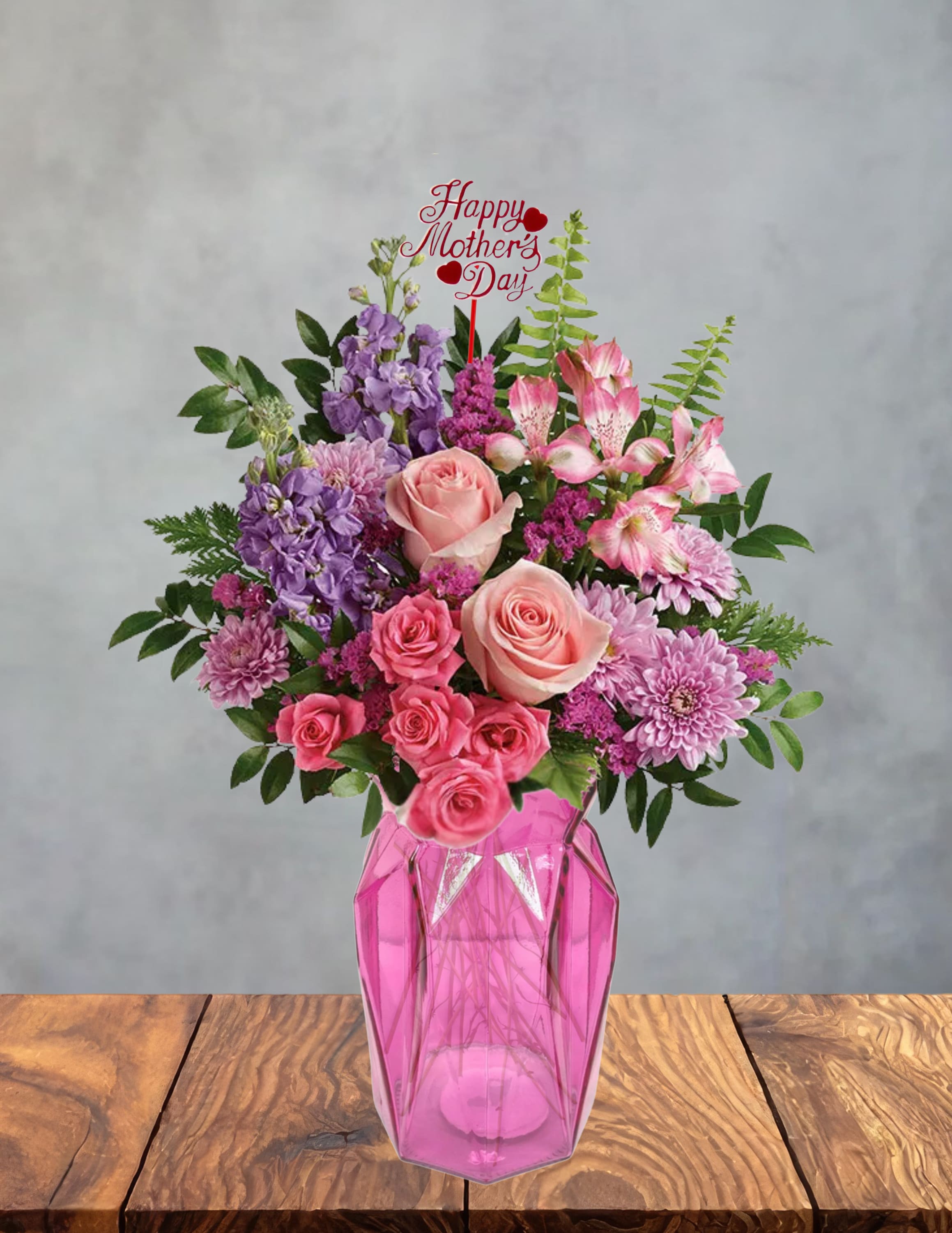 Moms garden glow - Soft pink roses and lavender stock burst from a faceted pink glass vase in a joyful Mother's Day bouquet designed to make her feel cherished. This flower arrangement (perfect as a Mother's Day gift or a springtime surprise) centers on feminine pinks and purples-magenta accents lift the palette so the person you're celebrating feels both seen and celebrated.  Densely arranged above the pink vase, the mix includes pink roses, purple stock, pink alstroemeria and lavender chrysanthemums with touches of green foliage for contrast. The rounded, full design reads lush from every angle and comes with a "Happy Mother's Day" pick tucked into the center for an extra celebratory touch.  Ideal for moms, grandmothers, or any woman who loves a soft, romantic color story, this arrangement brings a garden-fresh feel indoors and brightens mantels, dining tables, or bedside tables. Vase is included; keep flowers in fresh water, trim stems and change water every couple of days to extend bloom life.