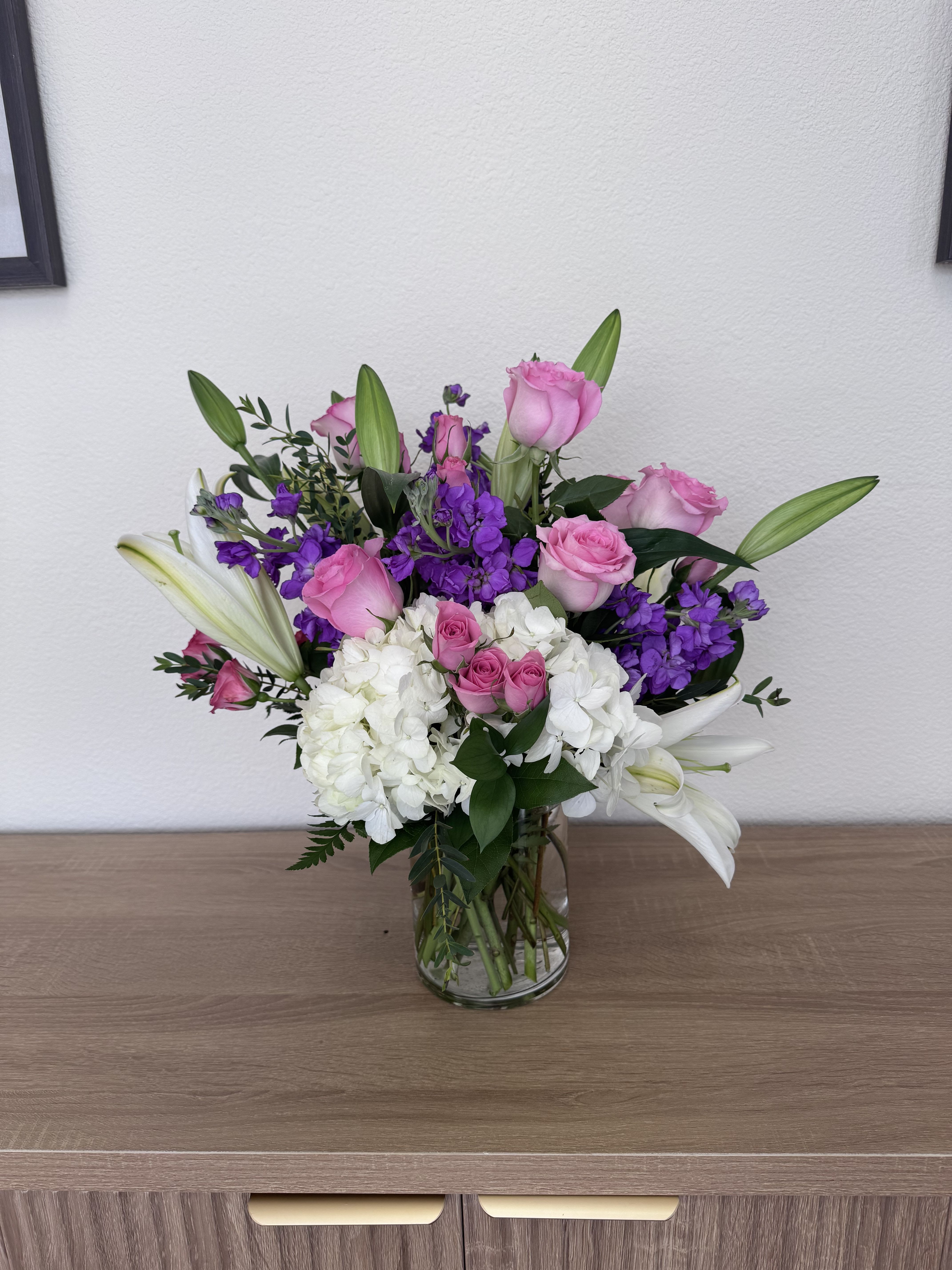 Simply Sweet for Mom - Soft pink roses and billowy white hydrangeas create a romantic foundation in this flower arrangement for Mother's Day, birthdays, or a thank-you surprise for Mom. The taller white lily buds rise above the rounded mass of blooms to give graceful height and movement, while purple stock threads through the composition for a bright, springlike contrast that feels both fresh and slightly formal.  Arranged in a clear glass vase, the design is full and layered-soft pink, crisp white, and vivid purple play against fresh green foliage for a balanced, textural look. The lilies add elegant vertical accents; the hydrangeas lend plush volume; and the roses bring classic, heartfelt charm. It's the kind of bouquet that makes the person you're thinking of feel truly seen and celebrated.