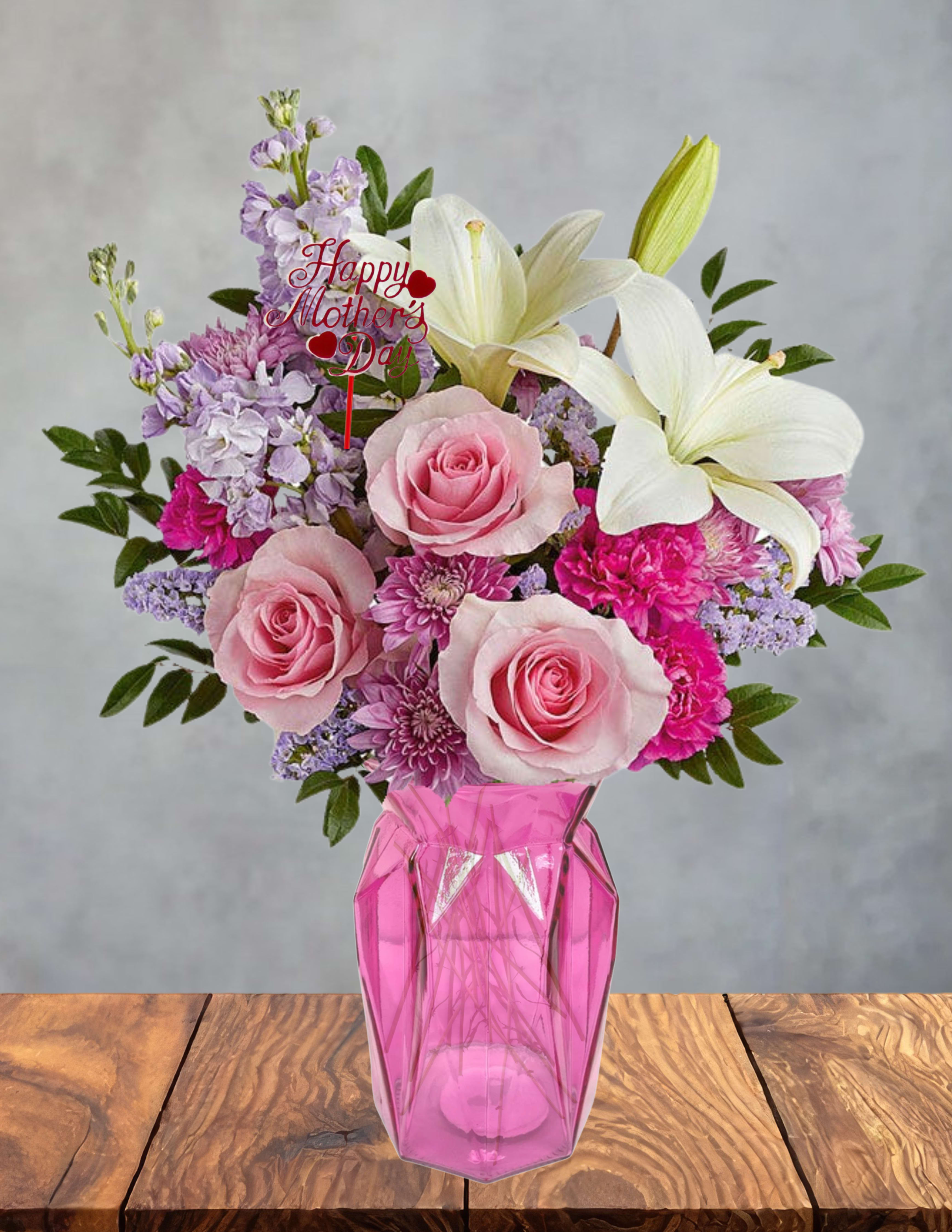 Her Day in Bloom   - Pale pink roses and fragrant white lilies mingle with lavender stock and bright magenta and purple filler blooms to create a soft, romantic Mother's Day flower arrangement that feels both feminine and joyful. As a Mother's Day bouquet, "Her Day in Bloom" is designed to make the person you're celebrating feel cherished and seen-its rounded, full composition reads as warm affection and thoughtful celebration.  The arrangement is gathered in a faceted pink glass vase that catches light and complements the pink, white, and purple palette. Three pale pink roses provide a gentle focal point while the white lilies add stature and scent; lavender stock and vivid magenta/purple fillers give texture and lively contrast. A small "Happy Mother's Day" pick is tucked near the top to clearly mark the occasion.  Perfect for moms, grandmothers, or any woman who deserves recognition, this bouquet suits a breakfast table, bedside, or entryway where it will brighten the room and invite compliments. Order with standard sizing as shown; keep lilies away from pets and recut stems with fresh water every few days to extend vase life.