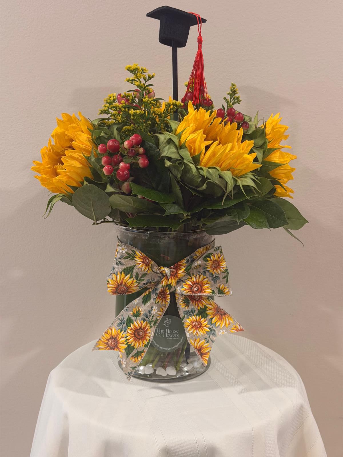 Sunflower Graduation Celebration Arrangement by The House of Flowers - Celebrate a proud milestone with our Sunflower Graduation Celebration Arrangement, a vibrant and joyful design that captures the spirit of success. This stunning arrangement features radiant sunflowers paired with fresh seasonal accents and lush greenery, beautifully arranged in a clear glass vase and finished with a charming sunflower ribbon.  Topped with a graduation cap detail, this piece is the perfect gift to honor achievements and brighten their special day.  Includes:  * Fresh sunflowers * Seasonal accent flowers and greenery * Clear glass vase with decorative ribbon * Graduation cap topper  Perfect for graduations of all levels and a meaningful way to say “Congratulations!”