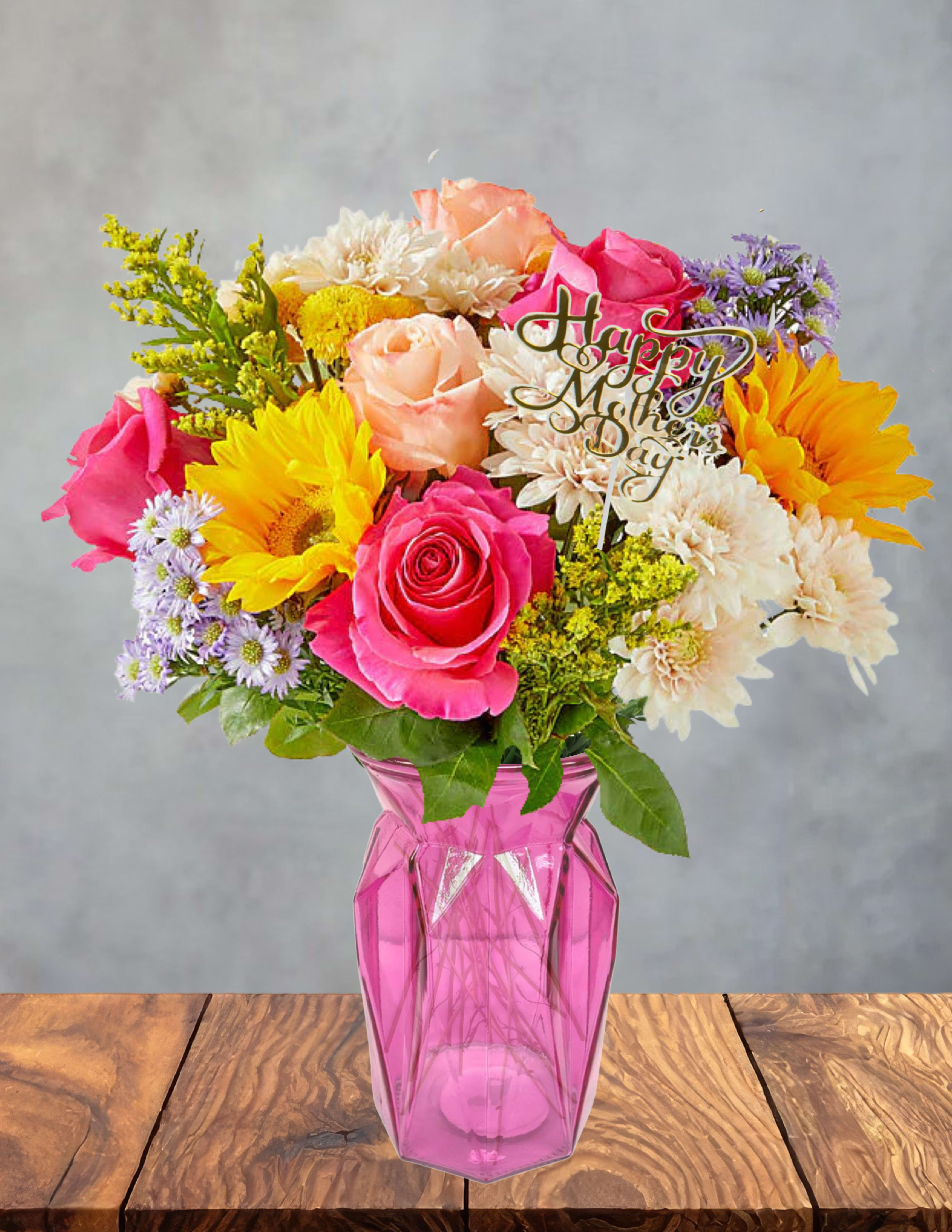 Petals of appreciation  - Bright pink and peach roses meet sunny yellow sunflowers in a lively Mother's Day bouquet that reads like a celebration in bloom. This flower arrangement for Mother's Day blends warm golds and cool lavenders - white chrysanthemums and small purple daisy-like blooms - so the person you're honoring will feel seen, cheered, and celebrated the moment they walk in the door.  Hand-gathered into a vivid pink glass vase, the mix of roses, sunflowers, daisies, and chrysanthemums creates a full, springtime silhouette with strong color contrast and a joyful texture: velvety rose petals, bold sunflower faces, and soft chrysanthemum clusters. A tasteful "Happy Mother's Day" pick is nestled at the center for an unmistakable gift moment; the vase provides a ready-to-display presentation that fits a mantel, table, or bedside.  Perfect for moms, grandmothers, or any woman you want to thank, this arrangement sends a warm, uplifting message without saying a word. Vase dimensions and care: keep stems in fresh water, trim 1/2" off stems every few days, and place away from direct heat or drafts to extend vase life.