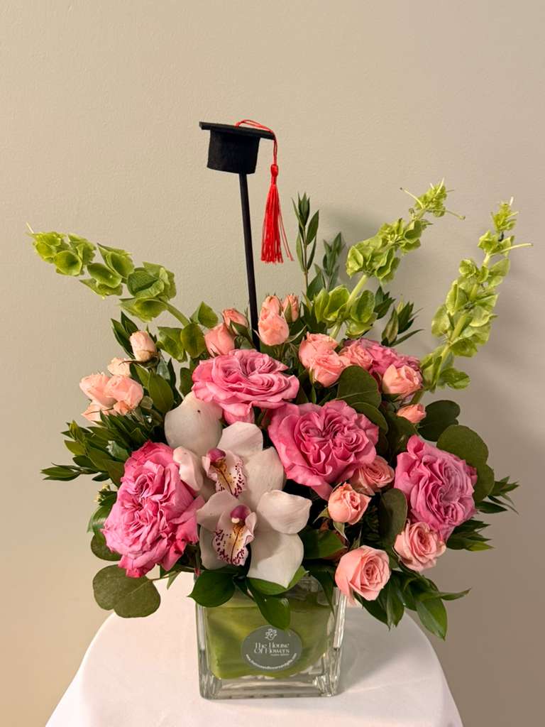 Blush Orchid Graduation by The House of Flowers - Celebrate a milestone in the most elegant way with our Blush Orchid Graduation Luxe Arrangement. This sophisticated design features premium garden-style roses in soft blush tones, delicately paired with spray roses, lush greenery, and a stunning orchid focal point that adds a refined, luxurious touch.  Presented in a modern glass vase and finished with a graduation cap detail, this arrangement is designed to impress and elevate any celebration.  Perfect for honoring graduates with style, grace, and unforgettable beauty.  Includes:  * Premium garden-style roses * Spray roses in blush tones * Orchid focal bloom * Seasonal greenery * Modern glass vase * Graduation cap topper