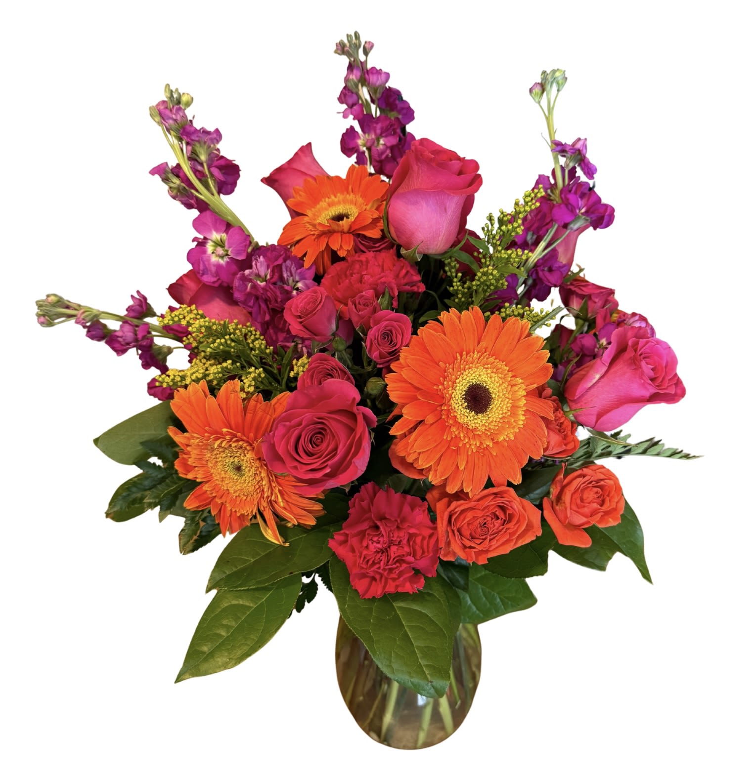 Fiesta for Mom - A burst of warm color that feels like a celebration: this flower arrangement is a bright, joyful bouquet perfect as a Mother's Day gift, birthday surprise, or just-because pick-me-up for the person you're thinking of. The lively mix of orange gerbera daisies and hot pink and coral roses creates an energetic, affectionate vibe that reads cheerful and romantic at once-she'll feel noticed and celebrated the moment she sees it.  The design pairs classic carnations, spray roses, and stock with magenta filler blooms and small yellow sprigs for contrast, all arranged upright in a clear glass vase. Several stems rise above the main cluster for a layered, airy silhouette; the palette of orange, pink, yellow, green, and purple gives plenty of visual texture and depth. The vase makes it ready to display on a table or mantel and keeps the arrangement looking fresh and arranged right out of the box.  This is the kind of bouquet that suits many happy moments-Mother's Day, congratulations, or a sunny birthday-while still feeling right on an ordinary day you want to brighten. For best results, place in fresh water away from direct heat and change the water every couple of days to enjoy the vivid colors longer.