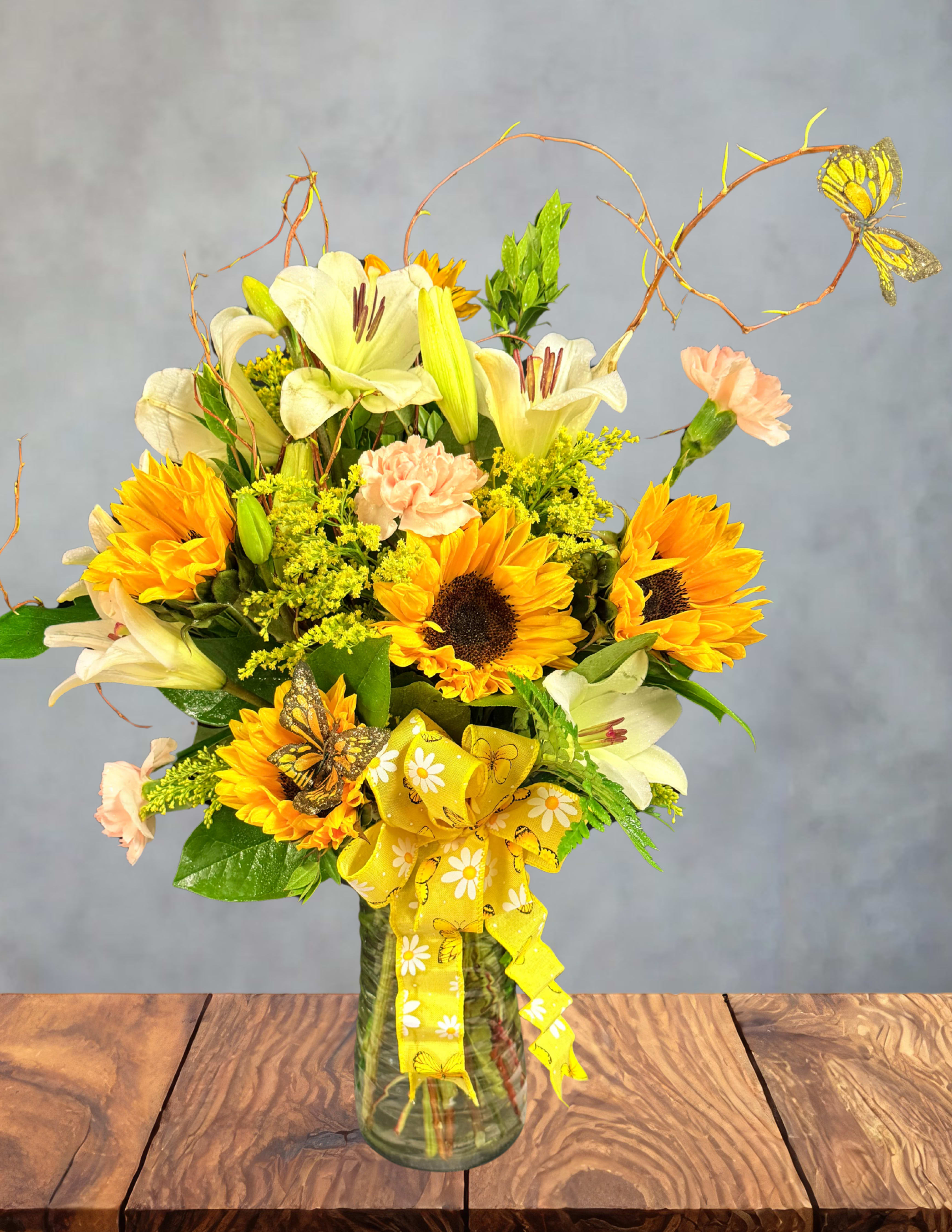 Wandering butterfly  - Bright sunflowers and white lilies dance together in this cheerful flower arrangement, making it an ideal birthday bouquet or everyday gift to brighten someone's day. The combination of bold yellow and crisp white with soft peach-pink carnations reads sunny and uplifting - the kind of floral surprise that tells the person you're thinking of them and will make them feel genuinely celebrated.  Set in a clear glass vase, the design pairs large sunflower heads with fragrant white lilies and velvety peach carnations, accented by small yellow filler blooms and curled twig accents for texture. A broad yellow decorative ribbon at the front gives it a finished, gift-ready look, while decorative butterfly picks add a playful, springtime touch. The arrangement's colors - yellow, white, pink, and green - create a lively, warm palette that fits cheerful seasonal occasions and casual celebrations alike.  Perfect for brightening a kitchen table, cheering a friend, or sending sunny birthday wishes, this arrangement arrives ready to display in the included vase. Change the water every few days and trim stems slightly on delivery to keep flowers looking fresh longer. Order early for same-week celebrations and let this wandering butterfly-themed bouquet bring a burst of warmth to the person you care about.