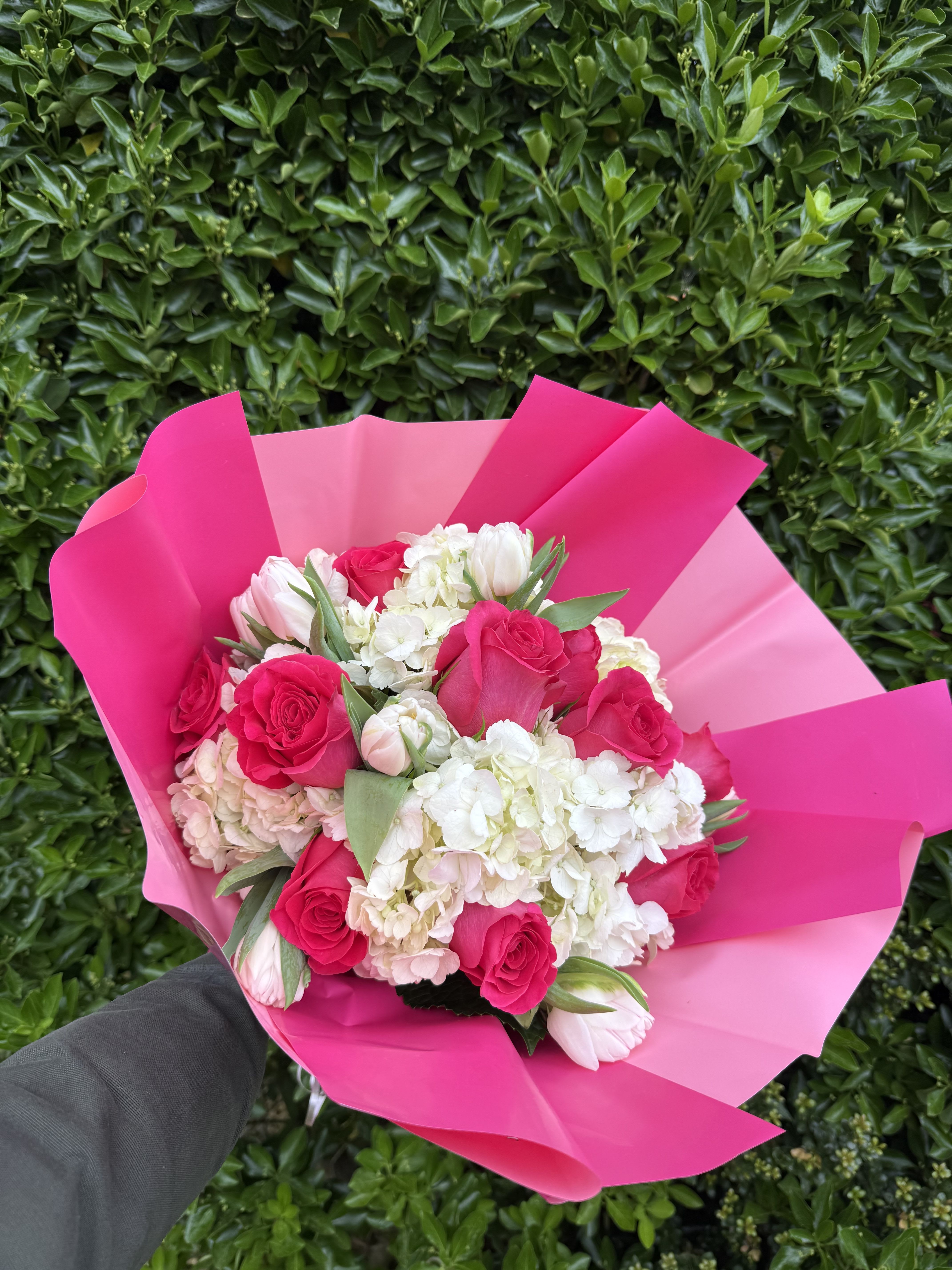 Mothers Day bouquet Wrap  - Vivid pink roses sit at the heart of this Mother's Day bouquet wrap, surrounded by clouds of white and blush hydrangeas and a few pale tulips for a soft, romantic contrast. As a flower arrangement for Mother's Day or a special birthday or anniversary, it reads as joyful and affectionate-the person you're thinking of will feel celebrated and cherished the moment they see the bright pink wrap and lush blooms.  The bouquet pairs rounded hydrangea clusters with the layered petals of roses and the graceful lines of tulips, creating a full, textured silhouette in white, pink, and green tones. Wrapped in a bold hot-pink floral paper, the presentation is cheerful and modern while keeping the overall feel tender and elegant. Designed to look abundant without being fussy, it makes a striking hand-delivered gift.  Order with confidence for Mom, a grandmother, or anyone who appreciates classic florals with a playful pop of color; this arrangement is ideal for in-person celebrations or to brighten someone's day from afar. To keep blooms fresh, place in clean water on arrival, trim stems at an angle, and change the water every other day-these simple steps help the hydrangeas, roses, and tulips last longer.