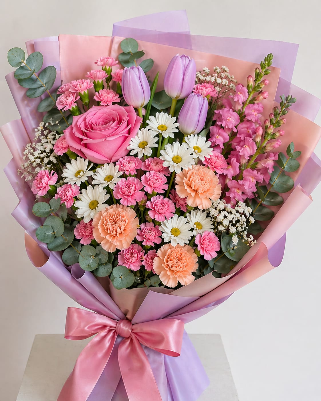 Pink Charm - A large pink rose takes center stage in this Pink Charm flower arrangement, a romantic and cheerful bouquet perfect for a birthday bouquet, anniversary roses alternative, or a thoughtful everyday gift to say "I'm thinking of you." Lavender tulips, white daisy blooms, and a mix of pink and peach carnations surround the focal rose, creating a soft spring palette that reads as both intimate and celebratory for the person you're honoring.  The stems are nestled with muted blue-green eucalyptus foliage and small filler flowers for texture, then wrapped in layered pink and lavender decorative paper and tied with a glossy pink satin ribbon for a polished finish. Expect a balanced, hand-tied size that feels substantial without being overwhelming-ideal for a bedside vase or a small dining table centerpiece.  Give this to someone who should feel genuinely seen: a friend celebrating a milestone, a partner on an anniversary, or a loved one who could use a bright, comforting surprise. Lightly rotate the vase every few days, refresh the water, and trim stems to extend freshness; available in the pictured color mix of pink, purple, white, and peach.