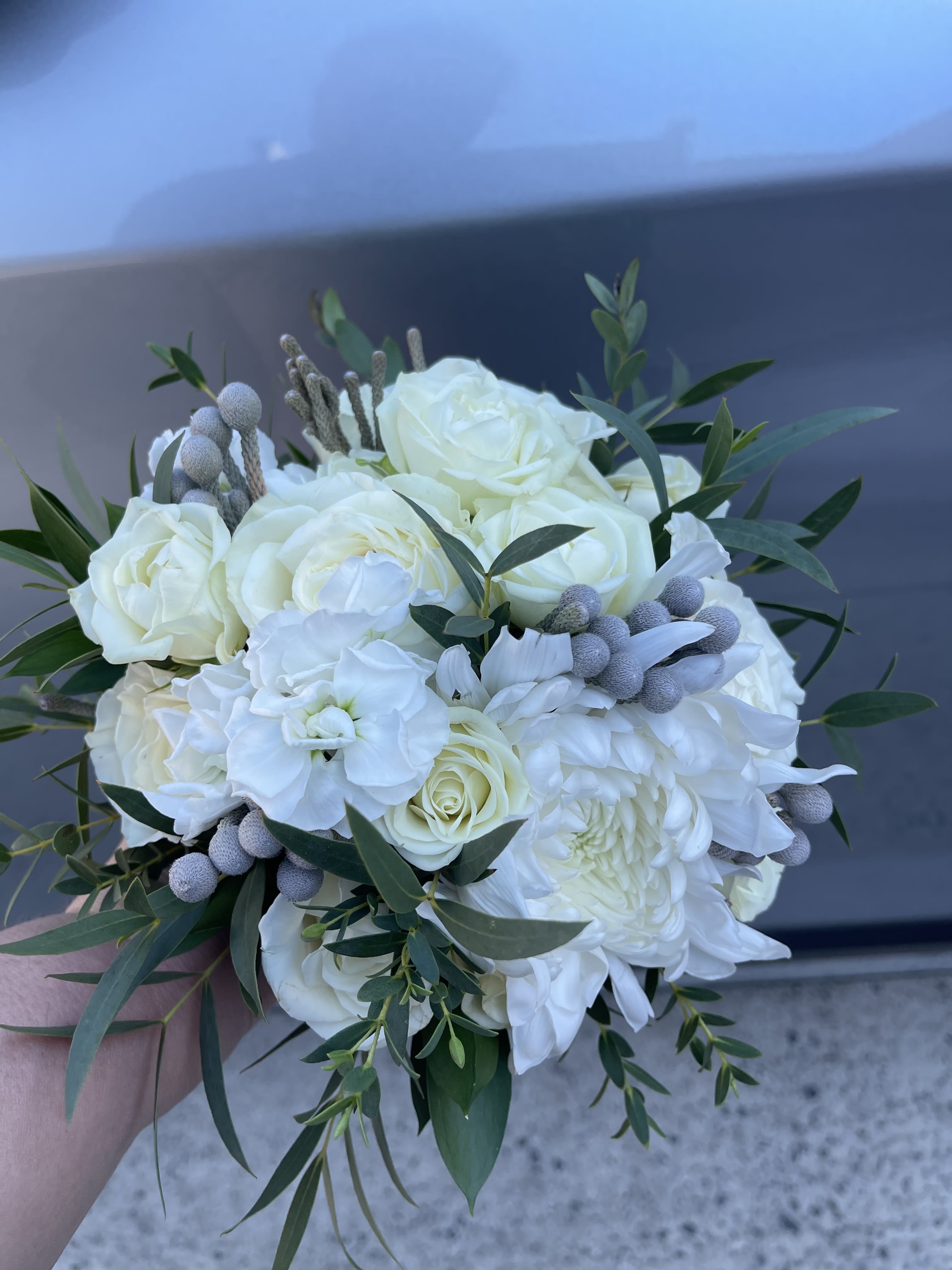Classic White Bridal Bouquet - A timeless all-white bridal bouquet featuring elegant roses, lush chrysanthemums, delicate blooms, and soft greenery accents.  Designed in a classic rounded shape, this bouquet offers a clean, sophisticated look that complements any wedding style — from modern to traditional.  Perfect for brides who love simplicity, elegance, and a refined aesthetic.  Each bouquet is handcrafted and may vary slightly while maintaining the same color palette and overall design.