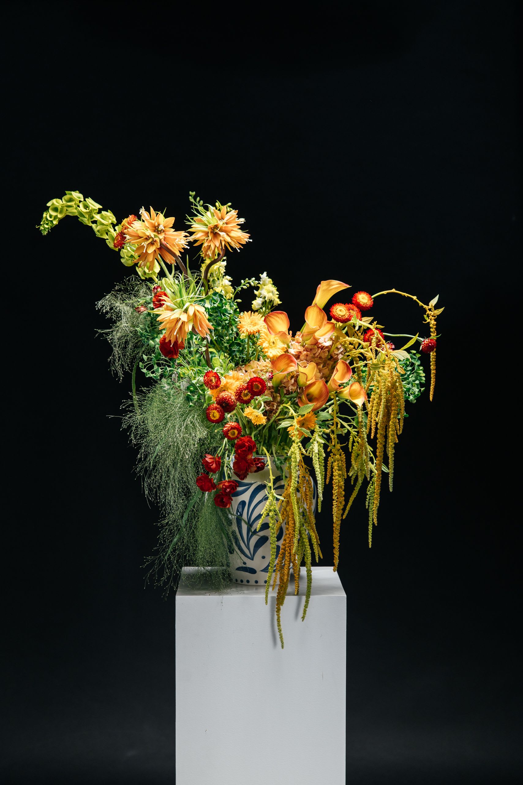 Zhar Ptitsa Vase Arrangement - Zhar Ptitsa is fire in motion. Inspired by the Slavic myth of the Firebird, this arrangement burns with оттенки of gold, amber, and deep orange—each stem placed to create movement, like flames caught mid-air. Sculptural lines rise and fall, while cascading textures flow downward like glowing feathers. It feels wild, untamed, and alive—never symmetrical, never predictable. This is not a safe choice. It’s for those who lead, not follow. For spaces that need energy. For moments that demand attention. A true statement piece—bold, artistic, and impossible to ignore.