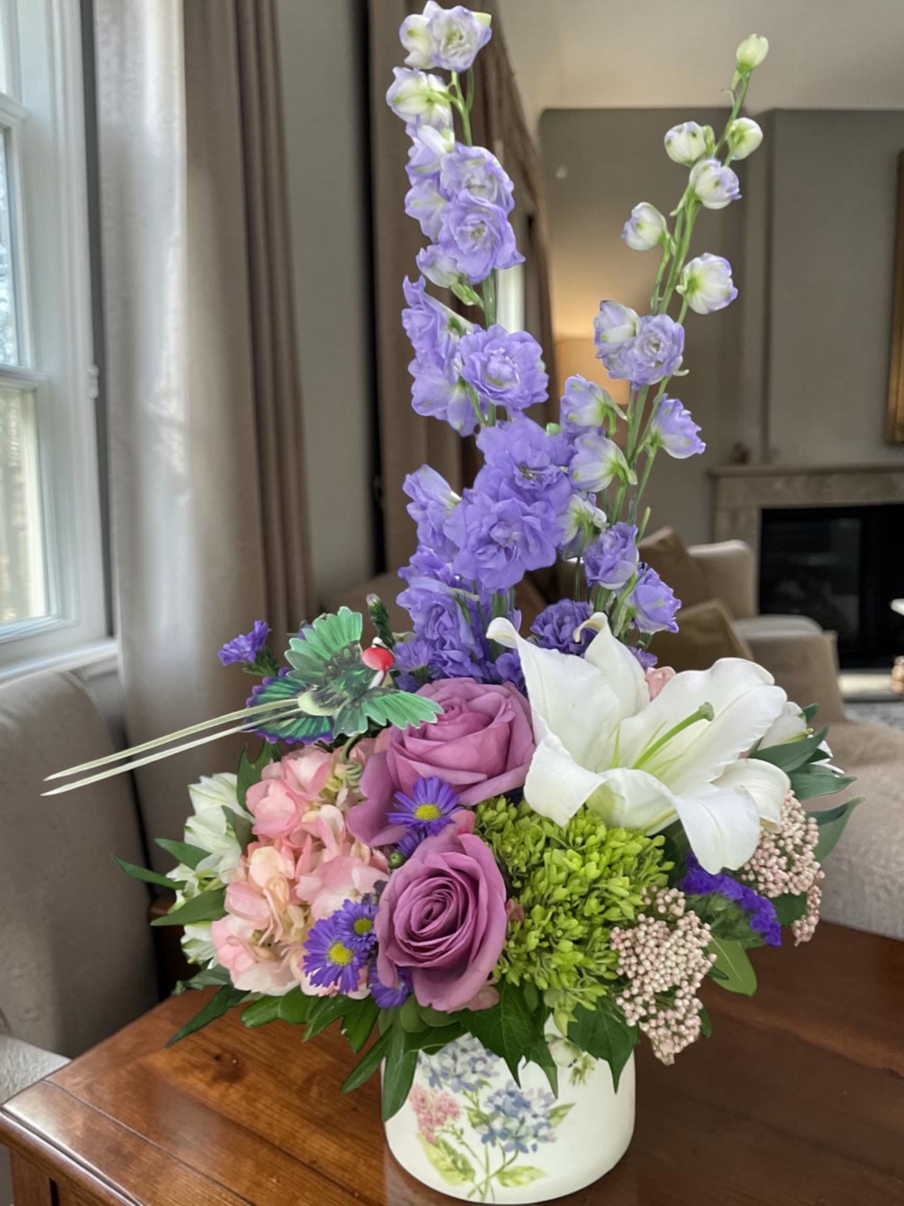 Fluttering Garden Mother’s Day Arrangement  - Celebrate Mother’s Day with the exclusive Fluttering Garden arrangement, a beautifully designed floral piece available only at Aspen Tower Florist in Greensburg, Indiana. This elegant, garden-inspired design features premium blooms including lavender roses, green and white hydrangea, a white oriental lily, soft alstroemeria, airy rice flower, and tall delphinium, all artfully arranged in a charming hummingbird keepsake container.  Designed in a soft palette of lavender, blush pink, fresh green, and crisp white, this arrangement captures the feeling of spring in full bloom. The graceful height of delphinium paired with a lush, full base creates a refined yet natural look, offering a balance of structure and softness that feels both timeless and fresh. The hummingbird container adds a meaningful touch, symbolizing joy, love, and lightness, making this more than a gift—it becomes a lasting keepsake.  Perfect for Mother’s Day flower delivery in Greensburg, Indiana, this arrangement is thoughtfully created to honor Mom with beauty and intention. Aspen Tower Florist proudly offers local delivery in Greensburg and surrounding areas, ensuring your flowers arrive fresh, on time, and ready to impress. We recommend ordering early for Mother’s Day, as this exclusive design is created with seasonal blooms and will be in high demand.