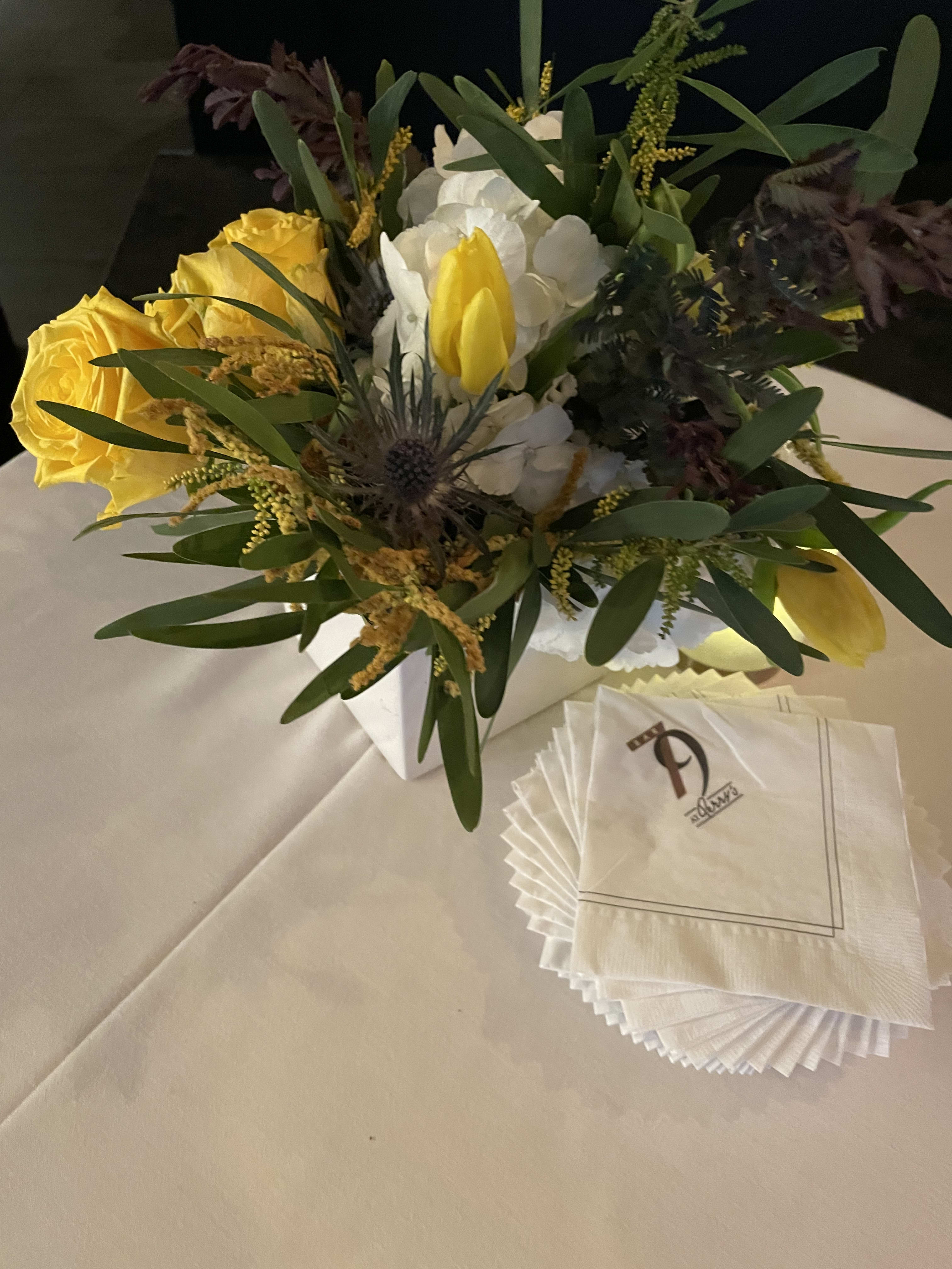 Yellow roses and white flowers arranged in a white box with napkins beside it.