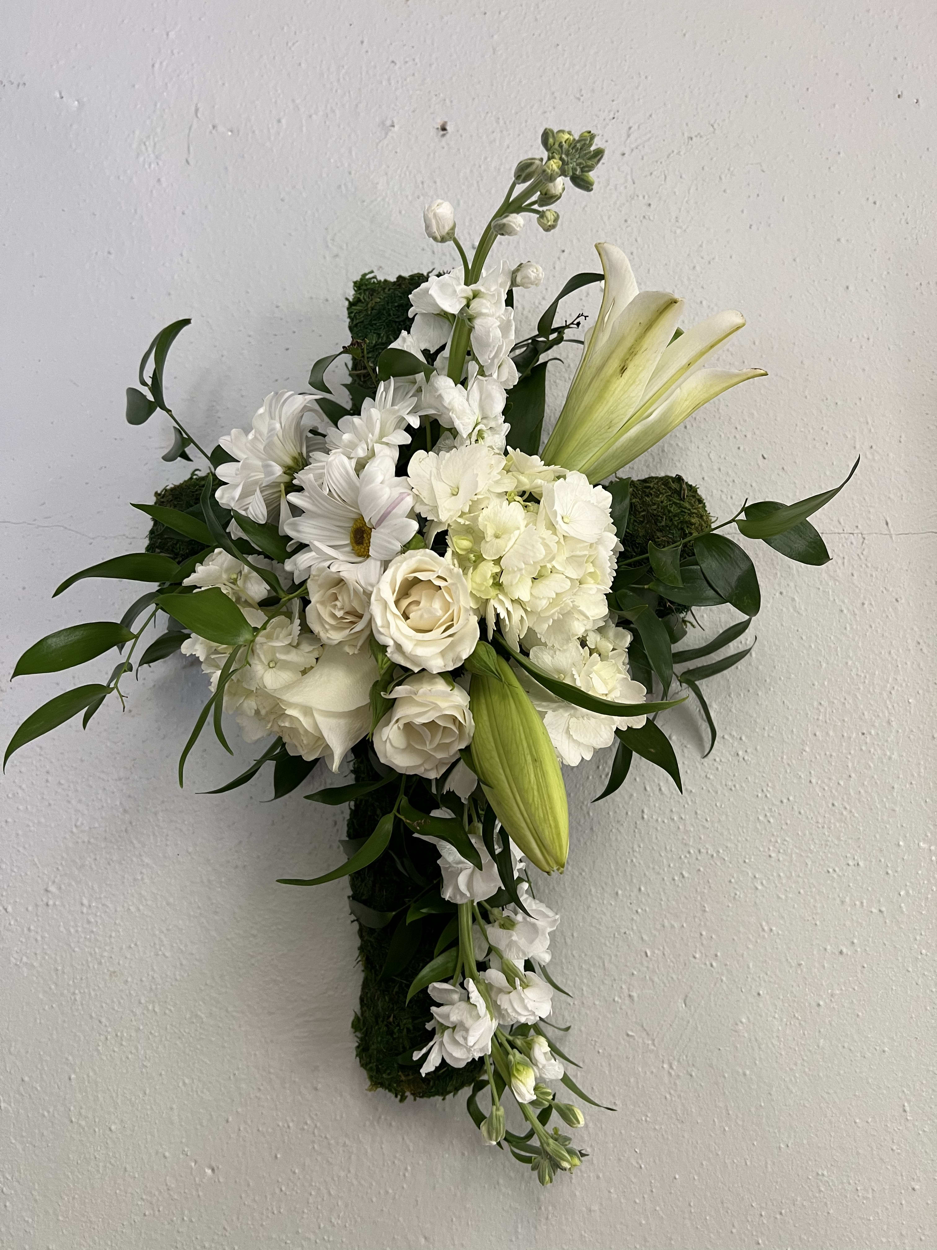 White lilies, roses, and daisies arranged on a moss-covered hanging cross.