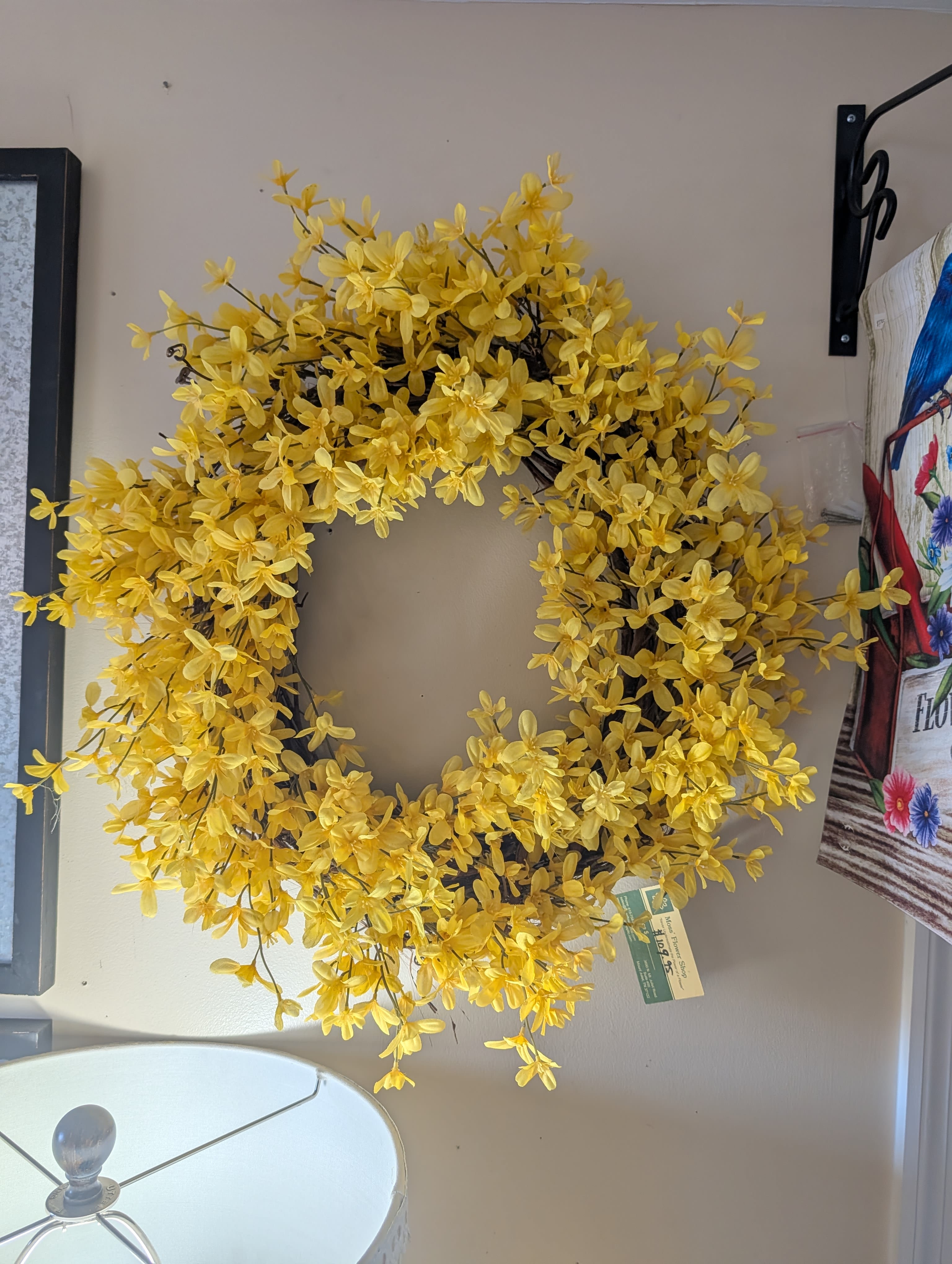Give Me Sunshine Permanent Botanical Wreath - A bright ring of cheerful yellow blooms greets the eye-this permanent botanical wreath is a wall-ready floral arrangement made to brighten entryways, mantels, or doors.   Dense clusters of small silk blossoms cover the full circular wreath form, creating a textured, almost cushion-like surface of saturated yellow punctuated by glimpses of dark, slender stems for natural contrast. The wreath's compact, full profile holds its shape whether hung on a door or propped on a wall hook, and because the blooms are permanent, it needs no watering-simply dust occasionally to keep the colors vivid.  Perfect for gifting to someone who loves a cheerful, low-maintenance touch, this piece says "thinking of you" or "happy spring" without fuss. Sized and styled for easy hanging, it arrives ready to display and will keep delivering sunny impact from season to season.