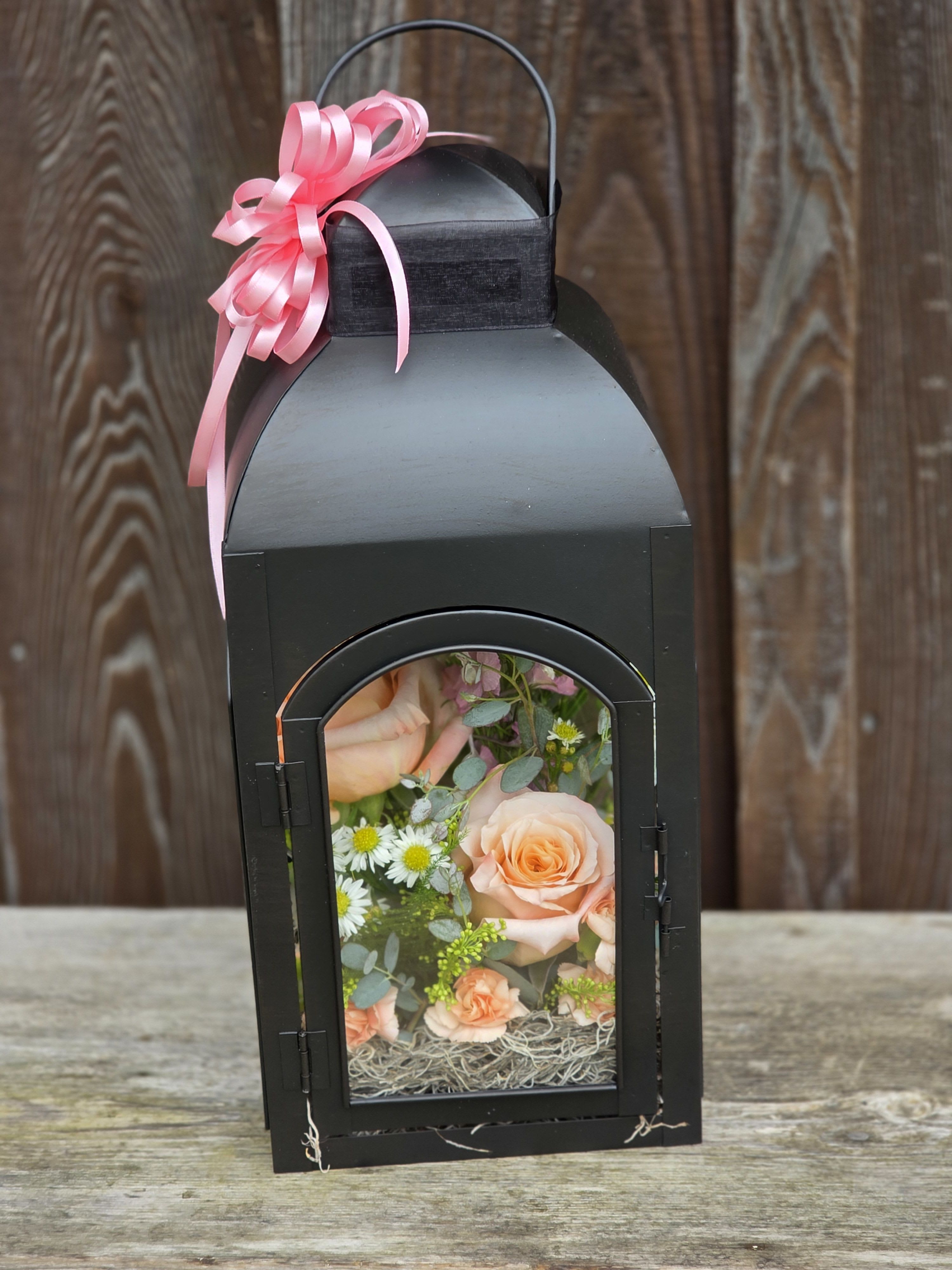 Black lantern with peach roses and white daisies inside, topped with a pink ribbon