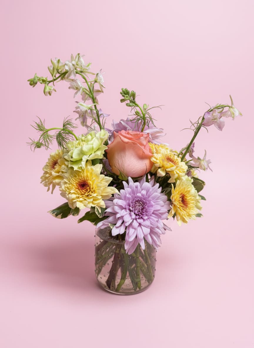 Morning Light  - Soft, radiant, and effortlessly charming, Morning Light captures the feeling of a slow, beautiful start to her day. Designed in a timeless mason jar, this arrangement blends coral roses in full, glowing bloom with the warm, velvety tones of crème brûlée cushions. Lavender cremones add airy texture and quiet elegance, while green carnations bring a fresh, lively contrast. Wisps of purple larkspur rise above it all, creating gentle movement and a graceful, gathered-from-the-garden feel.  Thoughtfully styled to feel both relaxed and refined, this piece is a sweet nod to simple moments made special—perfect for celebrating in a way that feels personal, bright, and full of love.  All flowers, floral colors, and containers are subject to change due to availability.