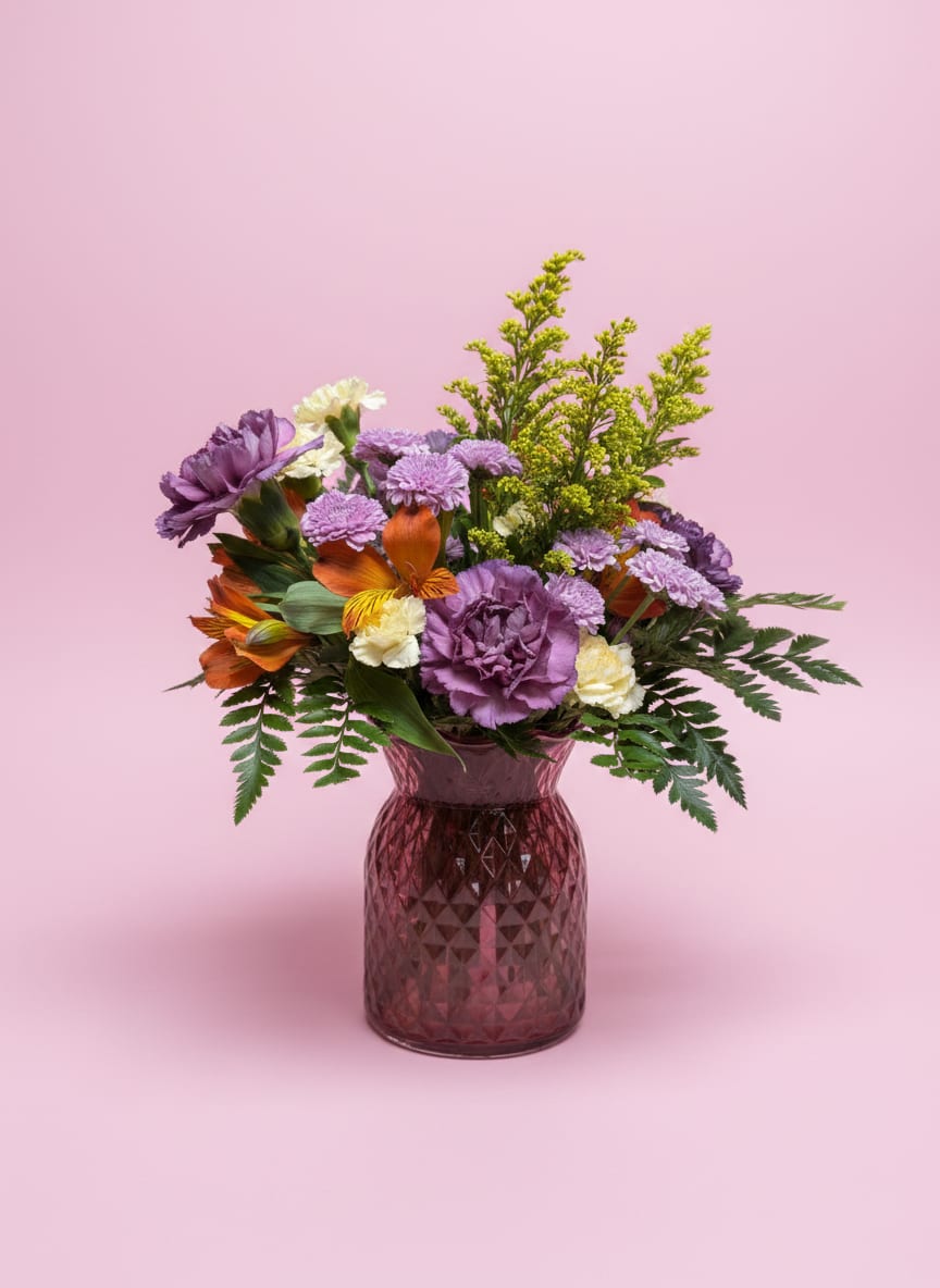 Gilded Grace  - Refined and radiant, Gilded Grace is designed to feel effortlessly elegant with a touch of modern glow. Arranged in a faceted diamond vase, this piece captures and reflects light beautifully, enhancing every detail within.  Metallic carnations bring a soft, luminous sheen, complemented by the gentle contrast of lavender buttons. Vibrant orange alstroemeria add natural movement and warmth, while yellow mini carnations and airy sprigs of solidago create a sunlit, layered finish.  Balanced, polished, and quietly striking, this arrangement is a graceful way to celebrate Mom—blending soft shimmer with fresh, joyful color in a design that feels both elevated and full of life.  All flowers, floral colors, and containers are subject to change due to availability.