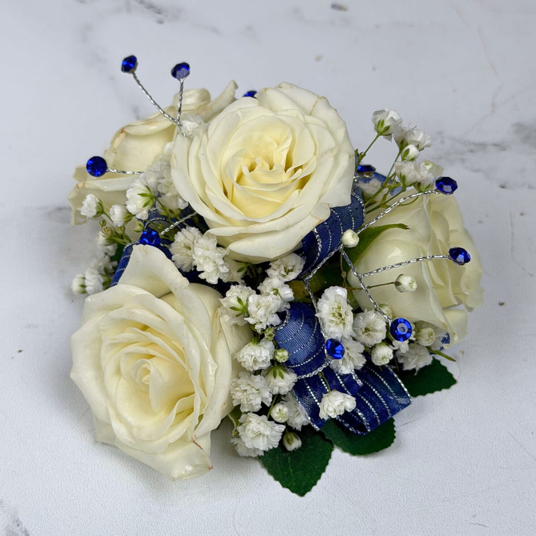 Sapphire 4 Rose Corsage - Cream-white roses gathered into a compact corsage give this Sapphire 4 Rose Corsage a refined look, perfect for prom. The cool contrast between ivory blooms and vivid blue accents creates a polished, celebratory impression that will make the person you're thinking of feel gracefully noticed on any formal occasion.  Four cream-white roses sit at the center, surrounded by clusters of tiny white filler blooms for soft texture. Wide blue ribbon loops provide a strong, elegant color pop while thin silver wire picks topped with bright blue gem-like rhinestones add sparkle and structure. The rounded, compact composition is small and wearable, designed to complement dresses and suits without overwhelming the outfit.  Choose this corsage for weddings, proms, anniversaries, or special ceremonies when you want someone to feel honored and seen. To keep it fresh, store it cool and avoid direct heat until the moment it's worn.