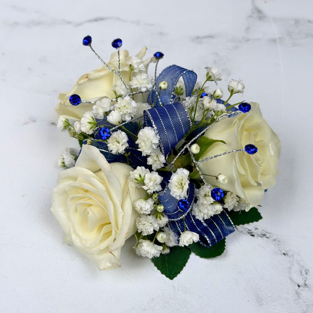 Sapphire 3 Rose Corsage - Cream-white roses gathered into a compact corsage give this Sapphire 3 Rose Corsage a refined look, perfect for prom. The cool contrast between ivory blooms and vivid blue accents creates a polished, celebratory impression that will make the person you're thinking of feel gracefully noticed on any formal occasion.  Three cream-white roses sit at the center, surrounded by clusters of tiny white filler blooms for soft texture. Wide blue ribbon loops provide a strong, elegant color pop while thin silver wire picks topped with bright blue gem-like rhinestones add sparkle and structure. The rounded, compact composition is small and wearable with an attached white lace wristlet, designed to complement dresses and suits without overwhelming the outfit.  Choose this corsage for weddings, proms, anniversaries, or special ceremonies when you want someone to feel honored and seen. To keep it fresh, store it cool and avoid direct heat until the moment it's worn.