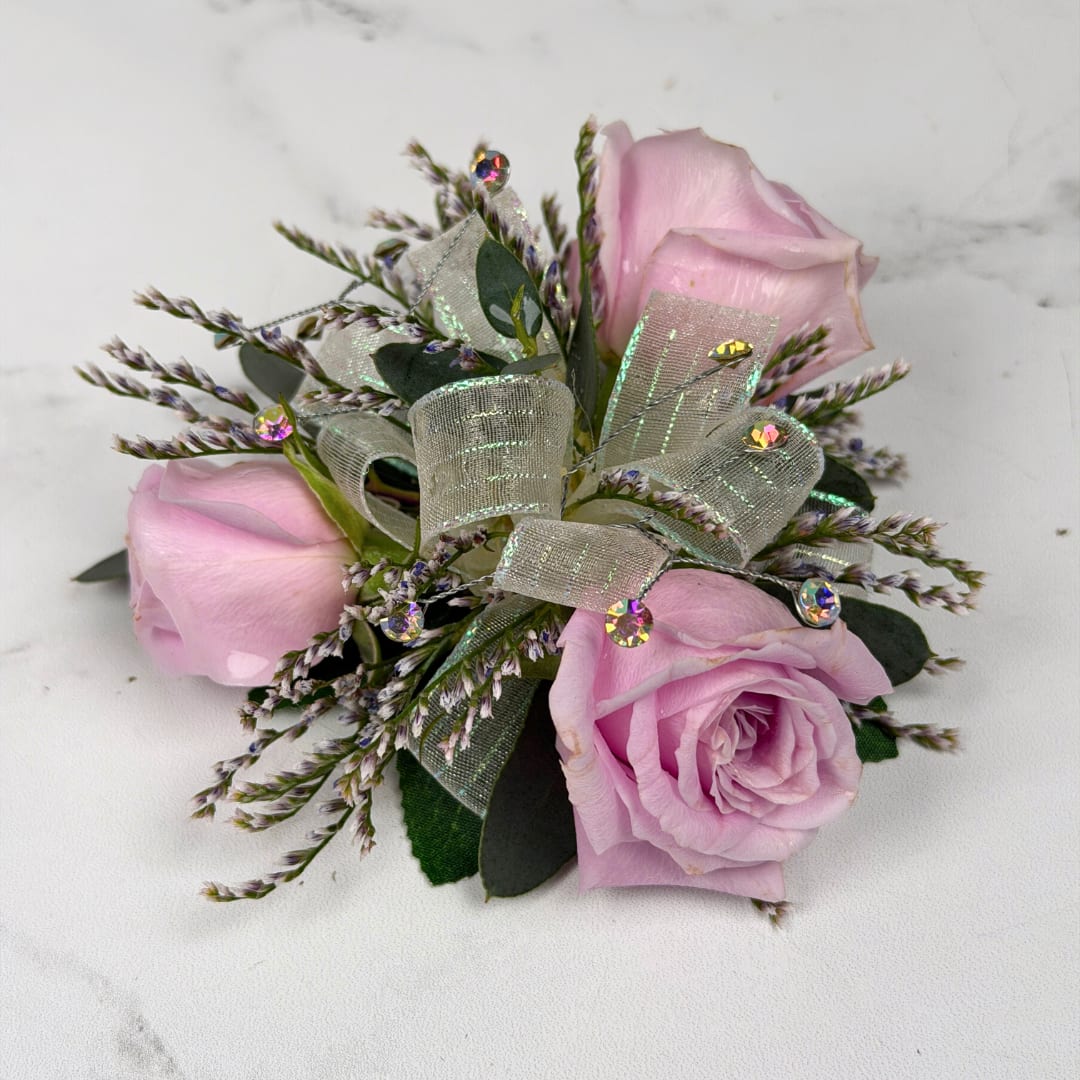 Amethyst 3 Rose Corsage - Three pale lavender roses form a delicate, wearable cluster in this Amethyst 3 Rose Corsage, perfect for prom. The soft lavender, white and green palette reads delicate and refined, the kind of piece that makes the person you're thinking of feel elegant and cherished on their big night.  The corsage pairs three compact roses with fine filler, finished with sheer iridescent ribbon loops and small iridescent rhinestone gems for a touch of sparkle. Hand-assembled to sit comfortably on the wrist with a white lace wristlet, it's small, neatly constructed, and designed to coordinate with formalwear and pastel palettes. Light misting and keeping it cool until wear helps maintain bloom freshness.