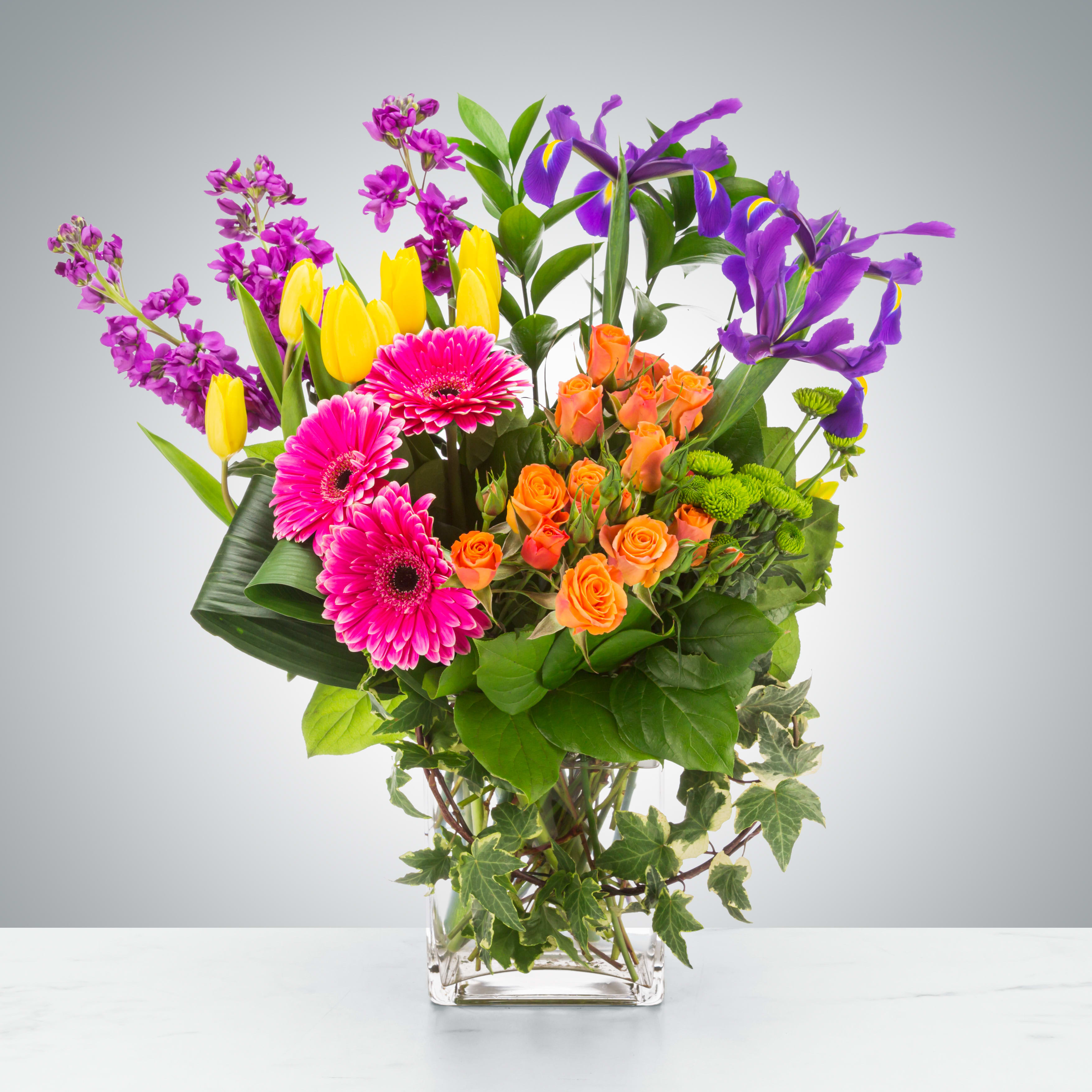 Mixed bouquet of pink gerberas, orange roses, yellow tulips, and purple irises in a glass vase