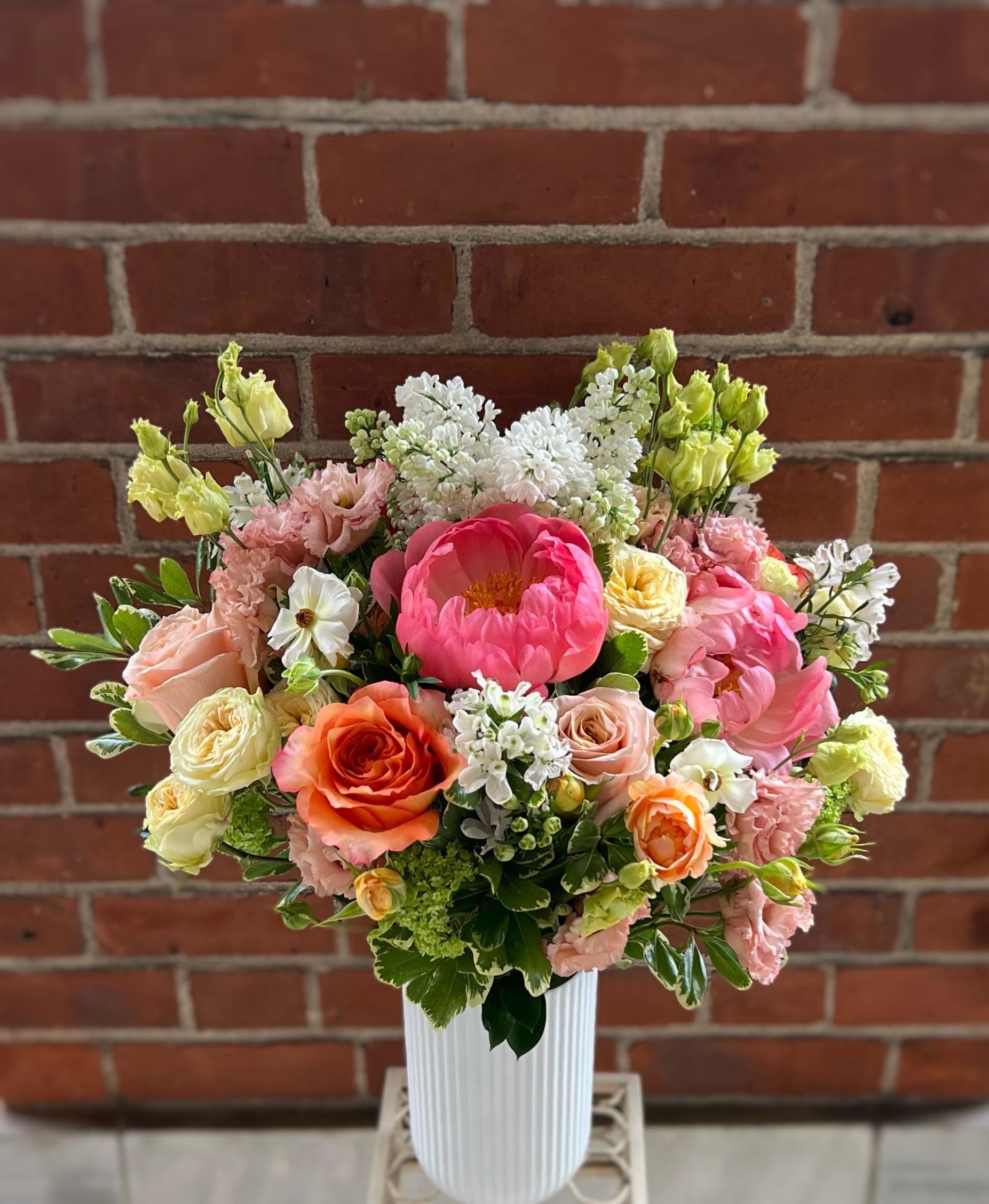 Tall arrangement of pink peonies, peach and orange roses, and white blooms in a white ribbed vase