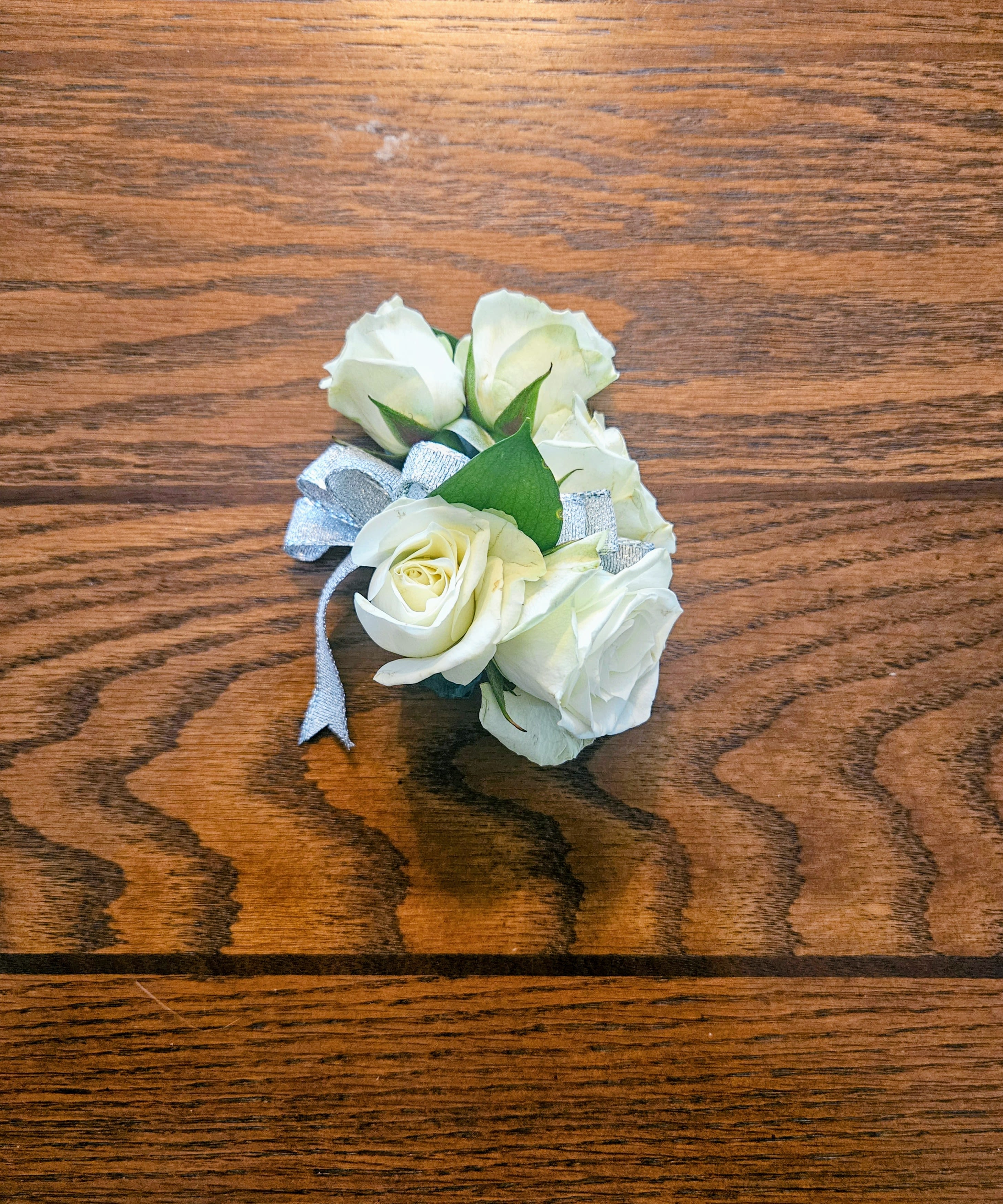 corsage - A cluster of white roses tied with a silver ribbon creates a timeless corsage perfect for weddings, proms, or other dressy events. This wrist corsage reads as soft and classic - the white blooms feel fresh and refined while the silver bow adds a subtle touch of sparkle that complements formal attire.  Each corsage is compact and wearable, made from several tightly clustered white roses with a few green leaves for contrast and finished with a neat ribbon bow. Options include sheer pastel bows in pink, lavender, or silver to coordinate with dresses or party palettes; the small scale keeps the look delicate and comfortable on the wrist.  Designed to make the person you're gifting feel celebrated and elegantly styled, these corsages pair especially well with bridal parties, prom dates, or honored guests. They're ready to wear upon delivery and should be kept cool and lightly misted until the event to preserve freshness.