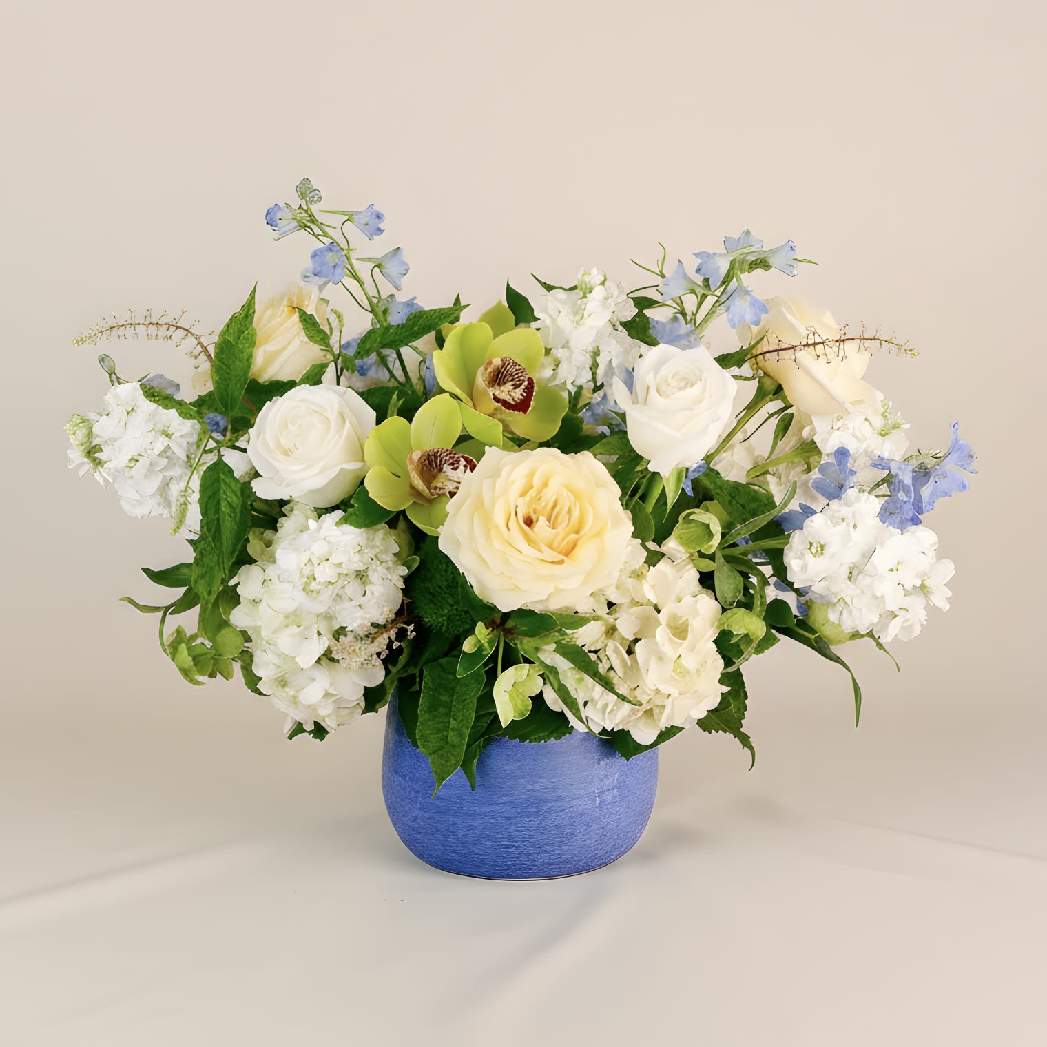 Santorini - A fresh and cheerful arrangement inspired by the colors of the Mediterranean. "Santorini" features cream and white roses, green cymbidium orchids, hellebores, light blue delphinium, white hydrangeas and more. Housed in a cerulean ceramic vase that evokes the iconic blues of Santorini’s rooftops, this design is perfect for brightening up any room! Arrangement approximately measures 20" tall and 25" wide with flowers.