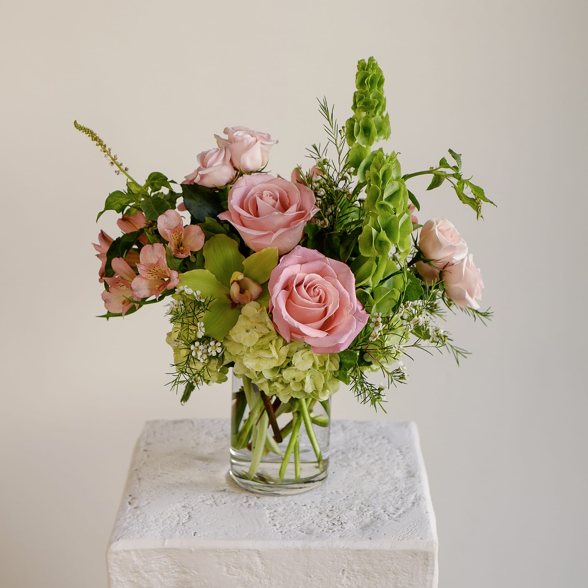 Hermosa - Lush pink roses and a green cymbidium orchid anchor this vibrant design, accented by architectural Bells of Ireland, green hydrangea, and soft pink alstroemeria. Arranged in a 4x6" clear glass cylinder, 'Hermosa' sits approximately 14 inches tall and wide, making it an ideal centerpiece or a refreshing accent for a console or desk. Every stem is hand-selected by our designers for exceptional quality and longevity. Local same-day delivery is available across West LA neighborhoods, ensuring the flowers arrive at peak freshness.