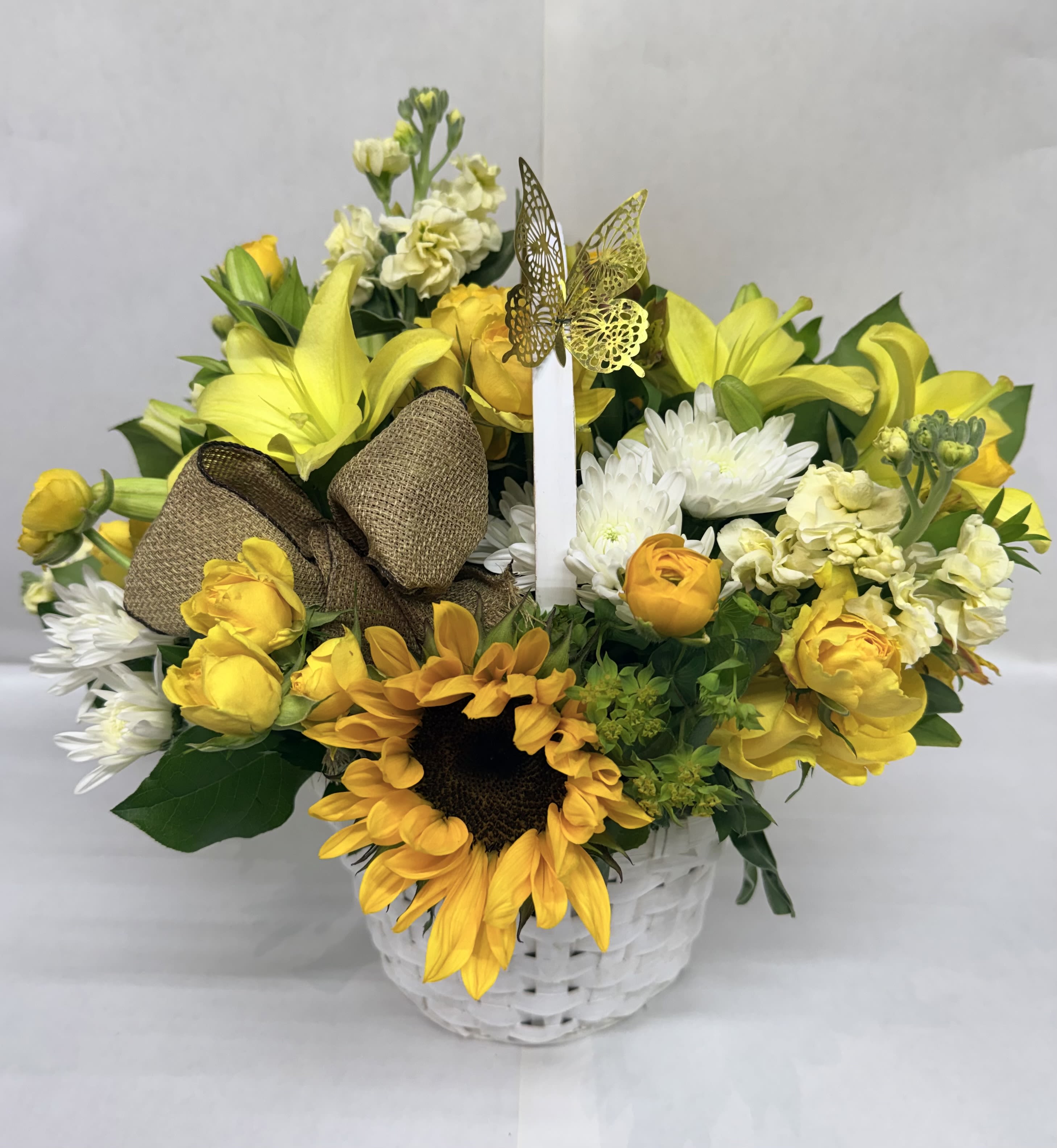 Sweetest Sunrise Bouquet - A bright sunflower anchors this cheerful flower arrangement, making it the perfect birthday bouquet or thank-you gift that says "you brighten my day." This flower arrangement for spring and everyday celebrations pairs sunflowers with yellow lilies, yellow roses, pale stock, and white chrysanthemums to create a sunny, uplifting display your recipient will notice the moment it arrives.  Arranged in a white woven basket, the composition balances bold yellow and crisp white blooms with fresh green foliage for a warm, seasonal look. A burlap bow and a decorative butterfly pick add a rustic, gift-like finish that feels hand-delivered and thoughtful. The mix of textures-from the sunflower's bold face to the soft petals of lilies and chrysanthemums-gives the bouquet a lively, natural presence.  Made to make someone feel genuinely celebrated, this arrangement is ideal for moms, friends, or coworkers who deserve a cheerful pick-me-up. It arrives ready to display in its basket; to keep blooms looking their best, refresh water for any floral foam daily and trim stems slightly if rearranging into a vase.