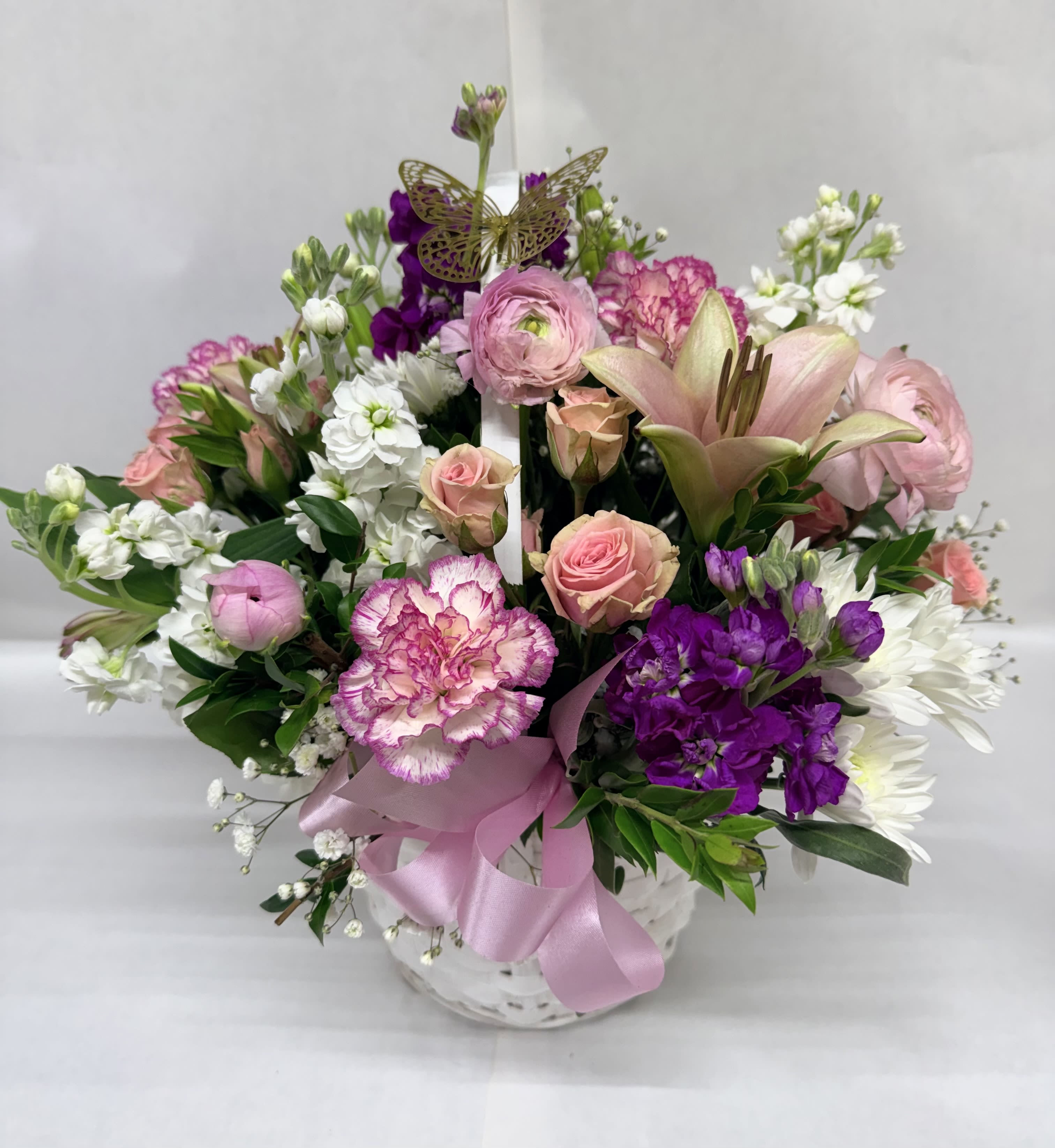 Be The Moment Basket  - A lush pastel basket that feels like a joyful moment captured - perfect as a birthday bouquet, anniversary gift, or "just because" surprise for someone you want to delight. This Be The Moment Basket presents a rounded, full arrangement in a white basket, combining pink, purple, and white blooms so the recipient immediately feels celebrated and cherished.  The design features fragrant lilies, soft roses, ruffled carnations, and clustered white stock, with small filler accents weaving texture and movement through the arrangement. Deeper purple blooms punctuate the soft pinks and whites for visual depth, while a pink satin ribbon tied at the base and a decorative butterfly pick rising above the spray add a whimsical, feminine touch.  Thoughtful for bedside tables, kitchen islands, or a reception desk, this basket sends the message that the person you're thinking of matters. Size and proportions are arranged to sit comfortably in a medium white handleless basket; light care-fresh water and a cool spot away from direct sun-will keep the blooms looking their best through the days they're meant to brighten.