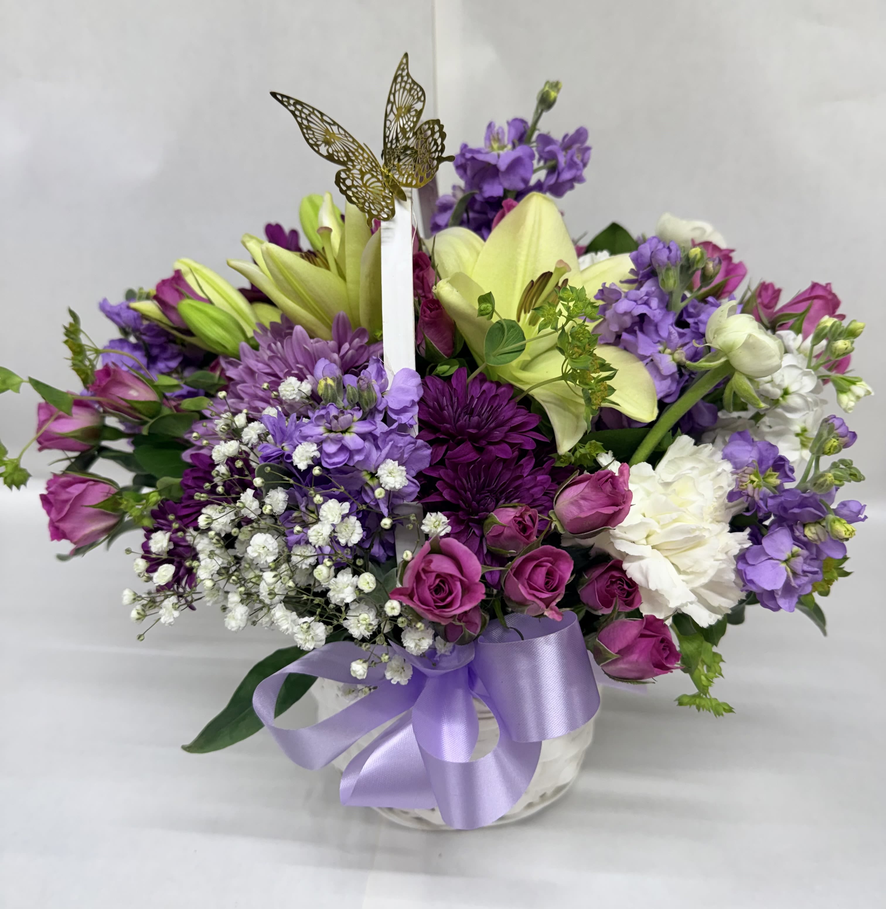 SWEET MOMENTS - A cheerful, romantic basket arrangement that feels like a springtime embrace-this flower arrangement for birthdays, anniversaries, or a thoughtful "just because" gift mixes purple, white, blooms to make the recipient feel truly celebrated. The bright yellow lilies rise near the center for instant impact, while pink roses and clusters of white filler flowers lend a soft, loving touch that reads as joyful and intimate.  Built in a rounded basket and finished with a lavender ribbon, the design layers purple stock and other purple blooms around crisp white flowers for contrast and texture. Daisies and additional filler blooms add friendly, airy texture, and a decorative butterfly pick hovers above the arrangement to give a whimsical, uplifting accent. The overall size is full and generous in the basket presentation, designed to sit beautifully on a table or display surface.  Perfect for marking birthdays, anniversaries, Mother's Day, or a springtime surprise, this arrangement sends a message of warmth and affection that will make the person you're thinking of feel seen and cherished. Lightly mist blooms with water and keep the arrangement in a cool spot away from direct sun to extend freshness; order early for delivery on the special day.