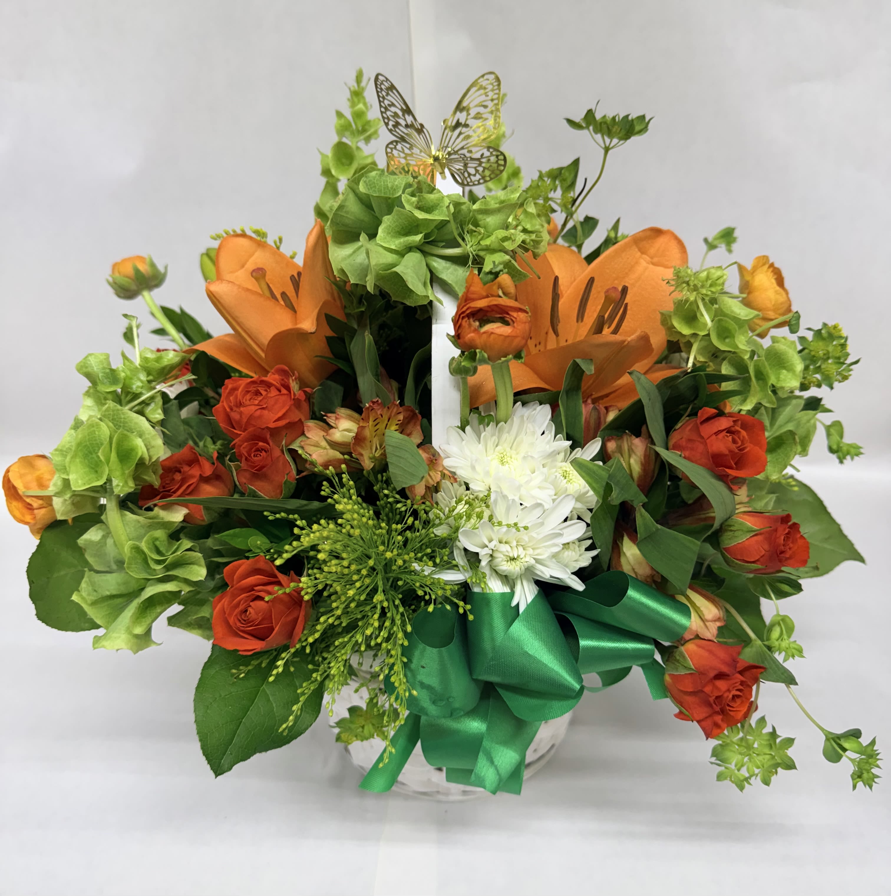 BLOOMING JOY BASKET - Bright orange lilies and velvety orange roses create a joyful, eye-catching bouquet perfect for an everyday celebration or a cheerful birthday arrangement. This flower arrangement pairs those warm tones with crisp white chrysanthemums and delicate alstroemeria to give the person you're thinking of a vibrant, layered display that reads as festive and heartwarming.  Arranged in a rounded basket , the composition uses green filler and foliage to build a full, textured silhouette while a large green satin ribbon at the base adds a polished, gift-ready finish. A decorative butterfly rises above the blooms for an added touch of whimsy and uplift. The mix of lilies, roses, chrysanthemums, and alstroemeria delivers varied shapes and scents that feel abundant without being fussy.  For the friend, family member, or colleague who deserves a sunny surprise, this basket-style arrangement says "celebrate today" and brightens any room. Designed to arrive ready-to-display, it's an easy choice for birthdays, thinking-of-you moments, or seasonal festivities; keep blooms in fresh water, out of direct heat, and they'll continue to bring that blooming joy for days.