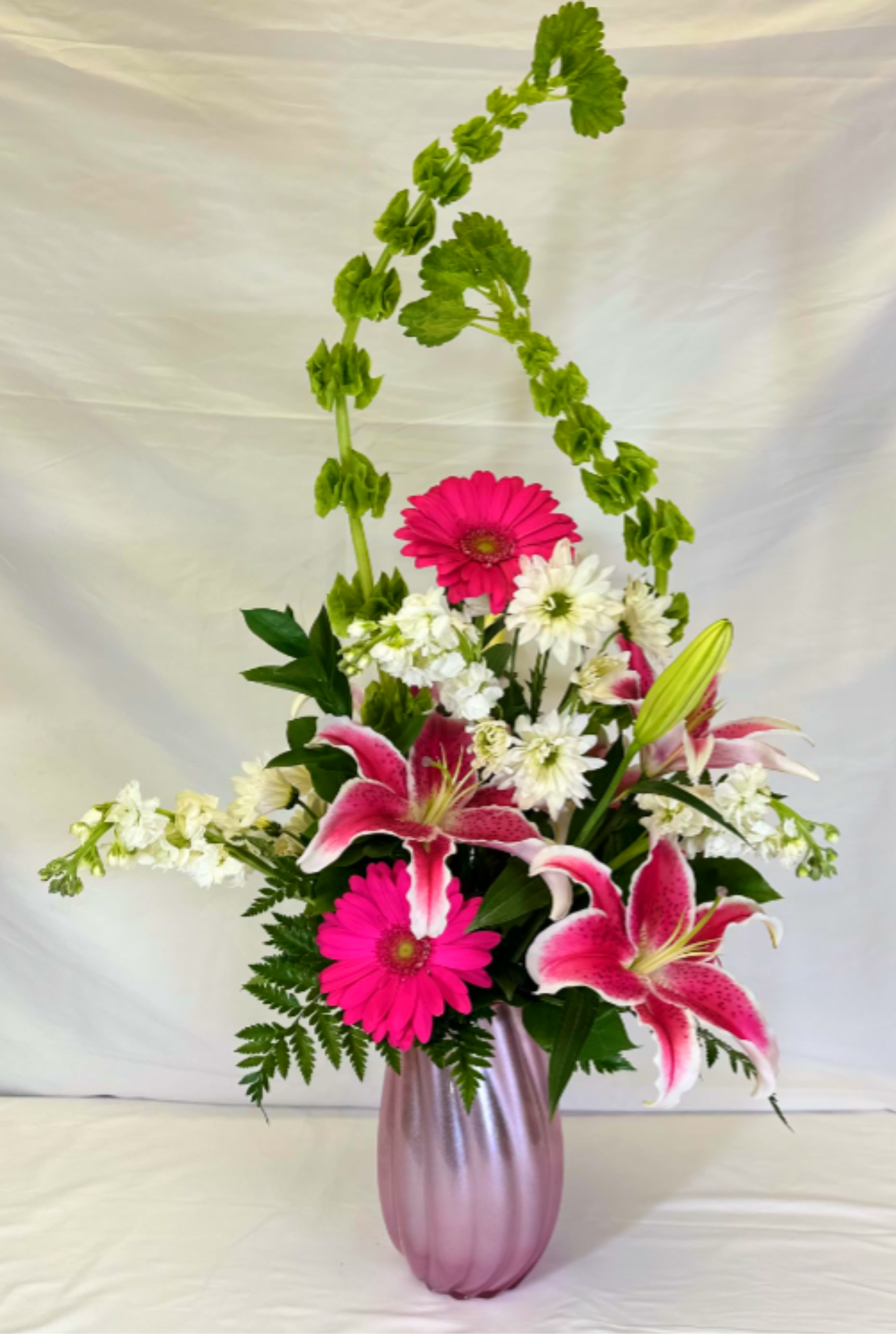 Tall pink and white arrangement with lilies and gerbera daisies in a pink vase with arching green bells of Ireland