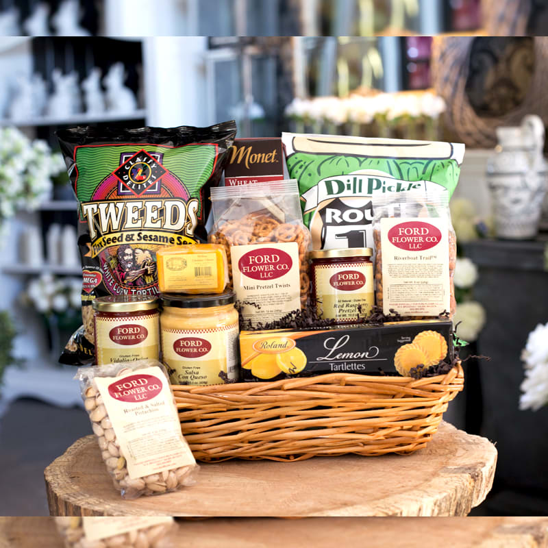Wicker gift basket filled with assorted packaged snacks and gourmet treats on a wooden surface.