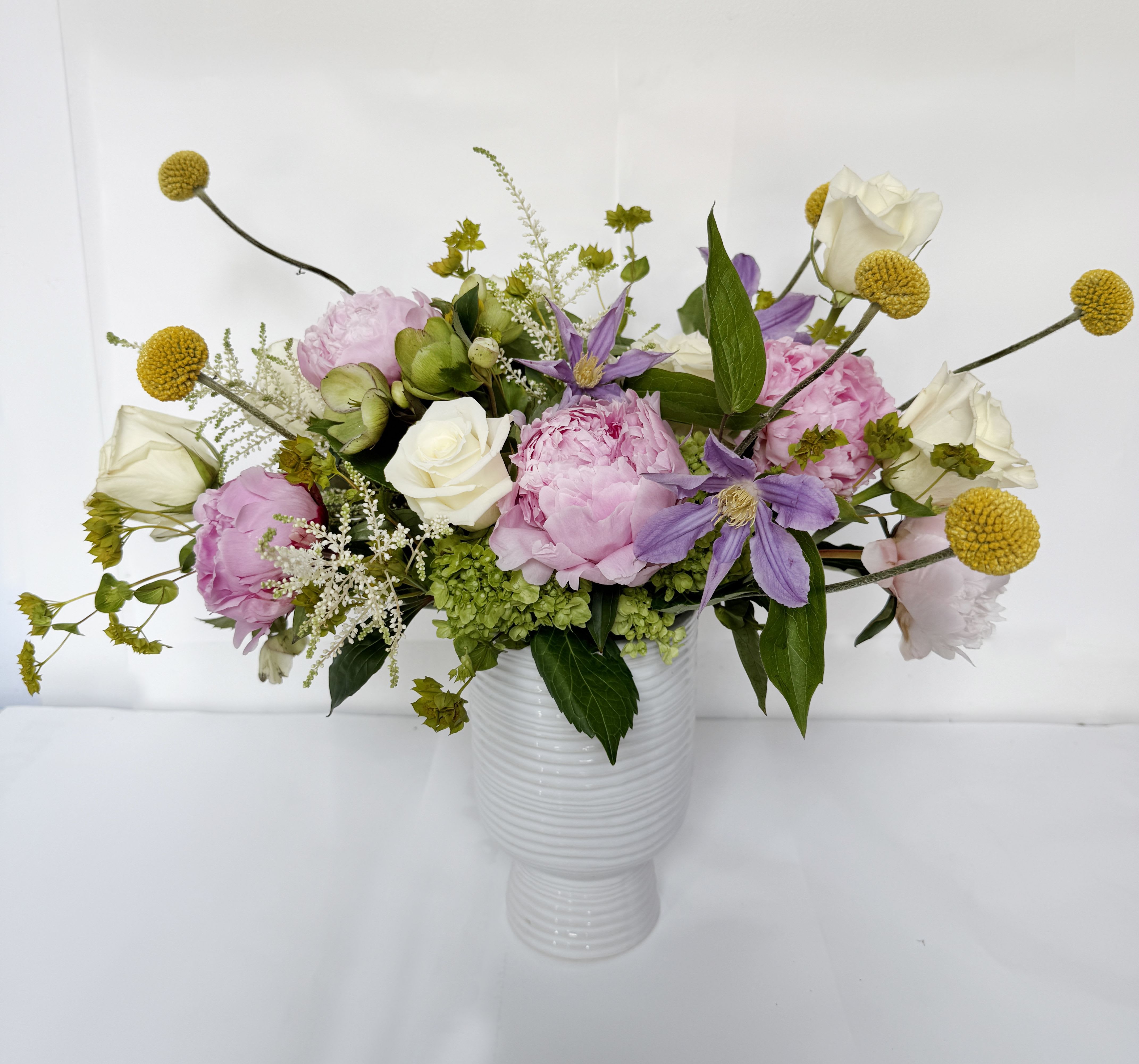 Stop at Monaco - This floral arrangement showcases a beautiful blend of white and pink hues, accented by a touch of vibrant yellow. It is thoughtfully arranged in a 5" by 7.75" white Monaco vase and includes elegant white roses, lush green hydrangea, charming pink peonies, and cheerful yellow craspedia.