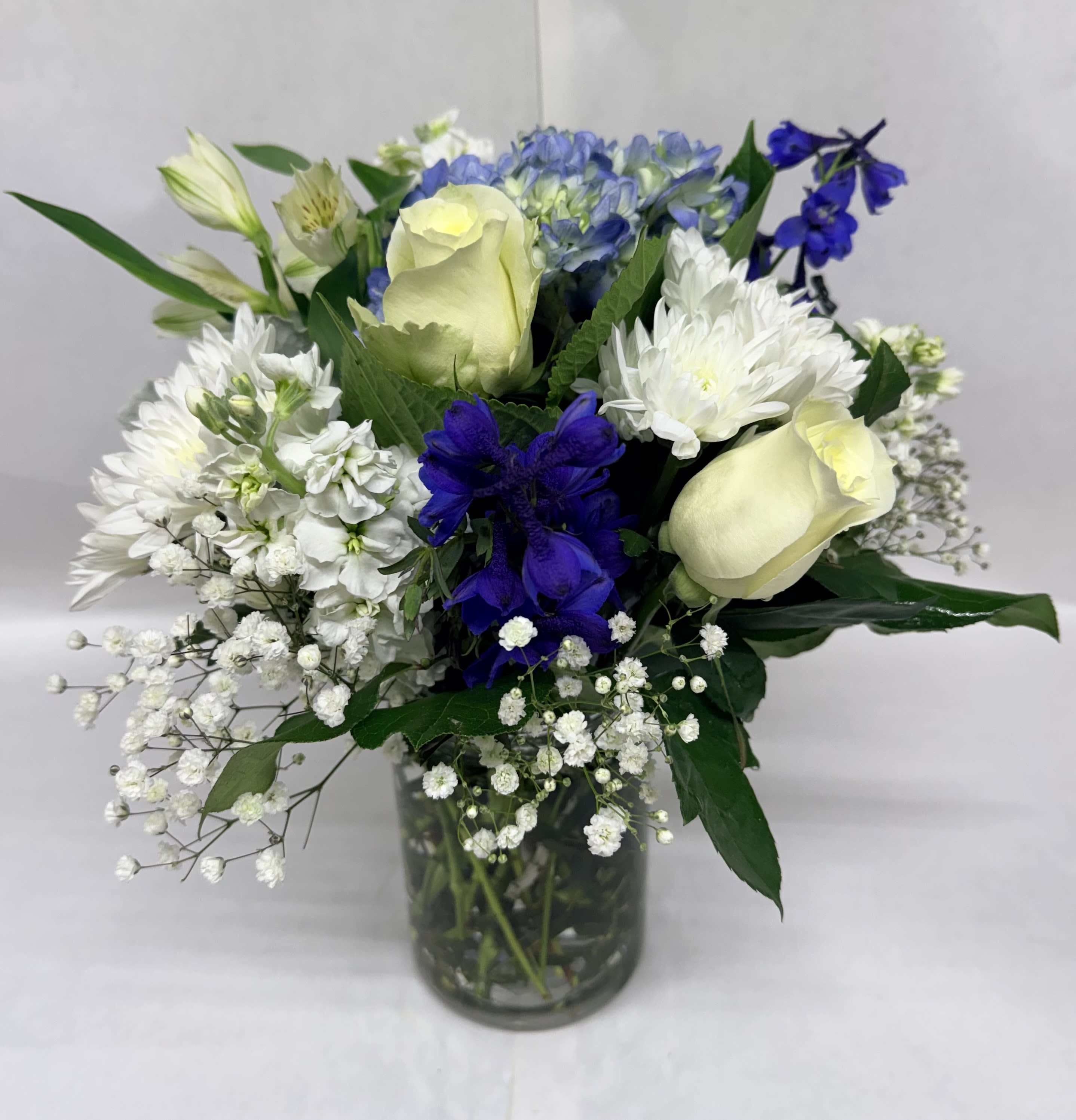 Clear Skies Bouquet - Pale white roses glow against a wash of cool blue hydrangea and crisp white pompom like blooms in this Clear Skies bouquet - a refreshing flower arrangement perfect for a birthday bouquet, a get-well gift, or a simple "thinking of you" surprise. The mix of soft blues, whites, and a touch of sunny yellow creates a calm, uplifting presence that will make the person you're thinking of feel noticed and comforted.  This full, rounded composition sits in a clear glass vase and pairs pale white roses with voluminous blue hydrangeas, white pompoms style blooms, alstroemeria accents, deep blue focal flowers, and airy baby's breath that softens the silhouette. The textures range from the lush, cloud-like hydrangea petals to the smooth, romantic rose blooms and the delicate sprays of filler, giving visual depth and a polished, gift-ready look.  Ideal for brightening a bedside table, office desk, or kitchen counter, this arrangement reads as both elegant and approachable - the kind of bouquet that calms and delights. Arrives arranged in a clear glass vase for immediate display; fresh cut-flower care (fresh water and a cool spot away from direct sun) will help the bouquet last longer.