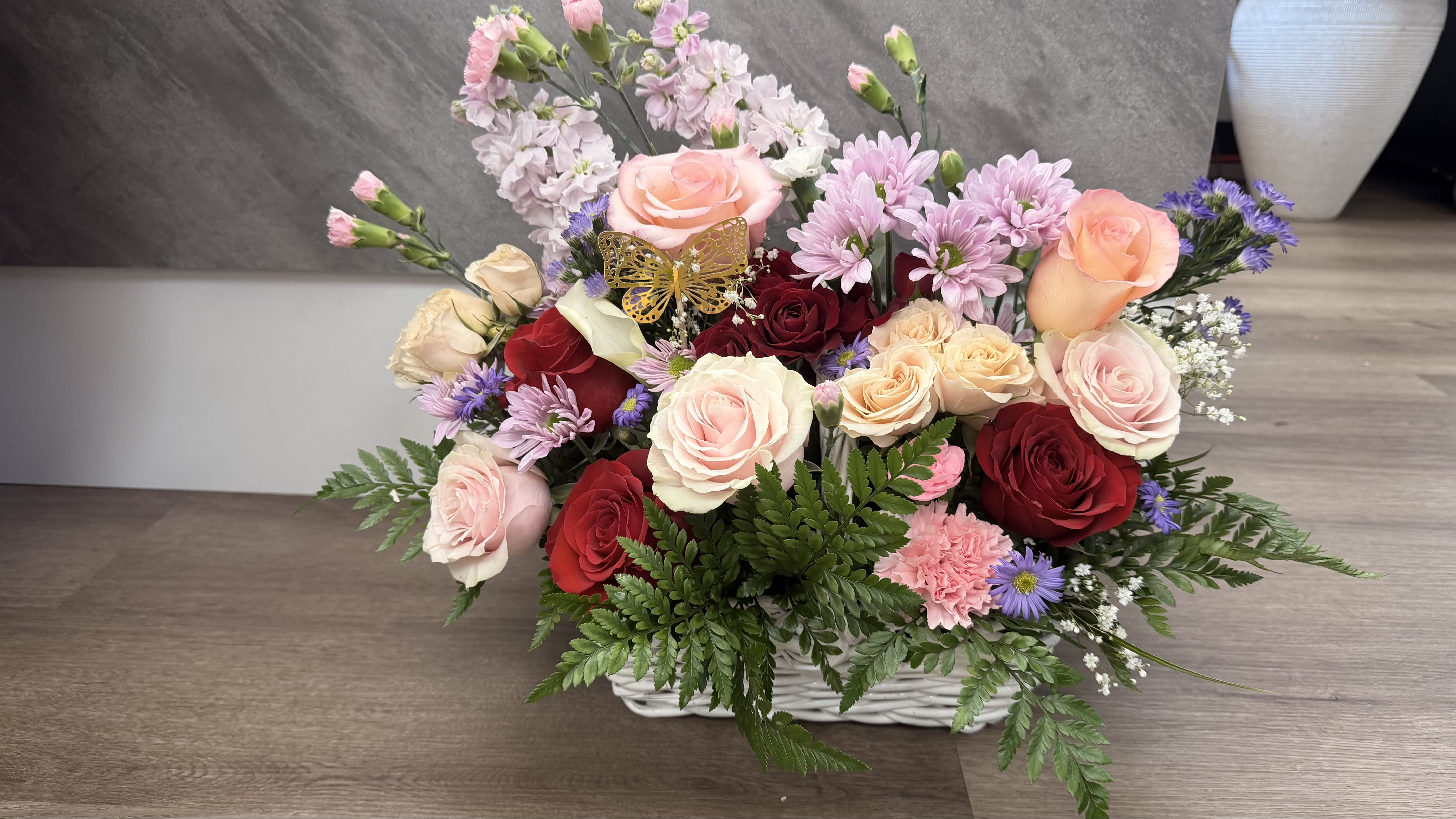 Pink basket - A lush bouquet of mixed roses and soft accents, this pink basket arrangement makes a romantic birthday bouquet or anniversary gift that will make the person you're thinking of feel truly cherished. The design reads as both cheerful and elegant-perfect for Mother's Day, a "thinking of you" surprise, or to brighten a get well room-while also fitting memorial, sympathy, or housewarming moments when a gentle, comforting touch is needed.  Dense clusters of blush, peach, cream, and deep red roses are layered with pink carnations and small purple daisy-like asters, all gathered low in a white wicker basket. Fern-like foliage spills outward to soften the silhouette, and a decorative butterfly pick near the center adds a whimsical, personal detail. The arrangement's low, rounded profile sits beautifully on tabletops or a mantle; sizing is compact yet full, making it an easy gift for birthdays, anniversaries, graduations, or as a thank you that reads as heartfelt and thoughtful.