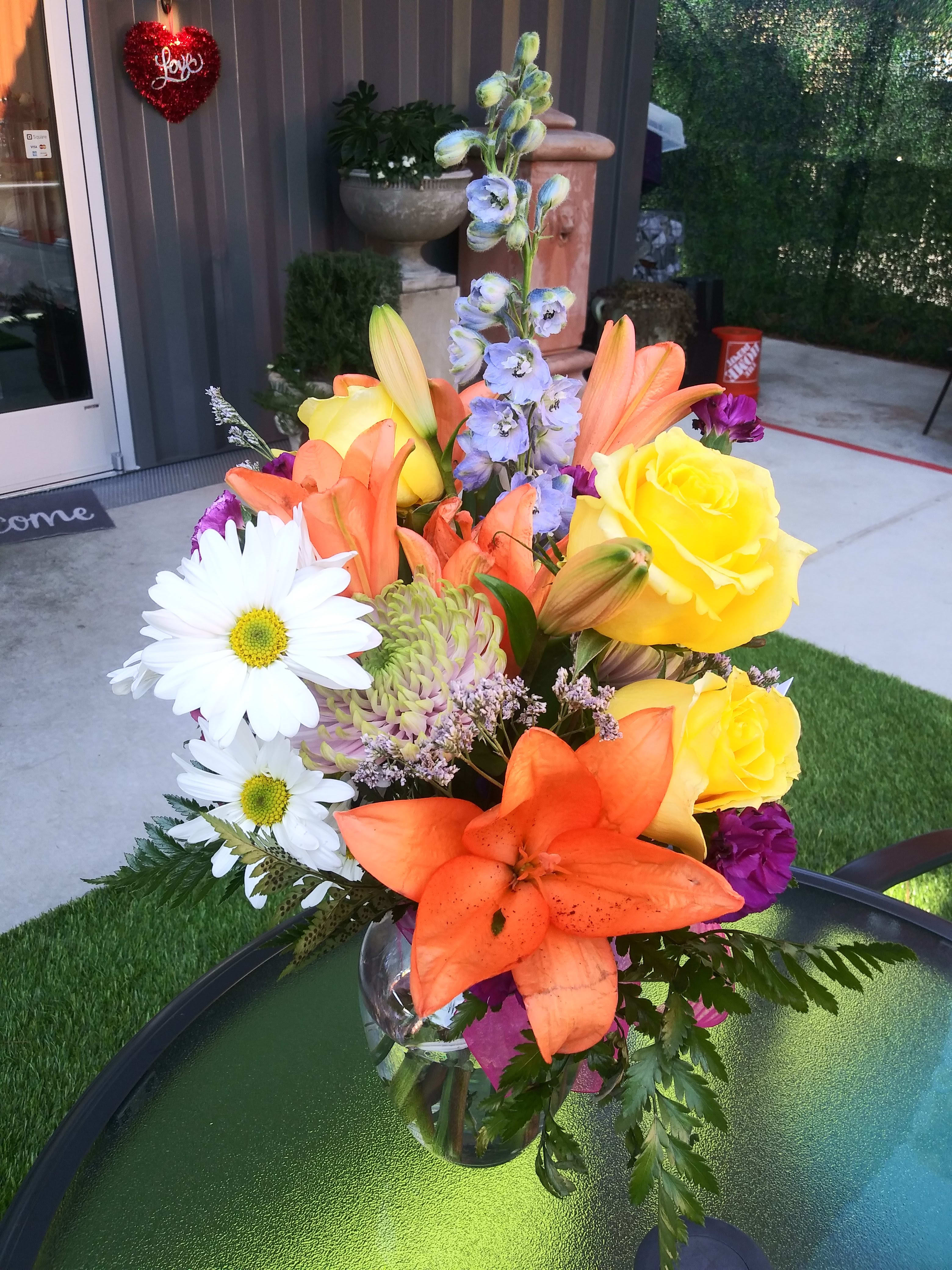 Bright mixed bouquet with lilies, roses, daisies, and purple accents in a glass vase