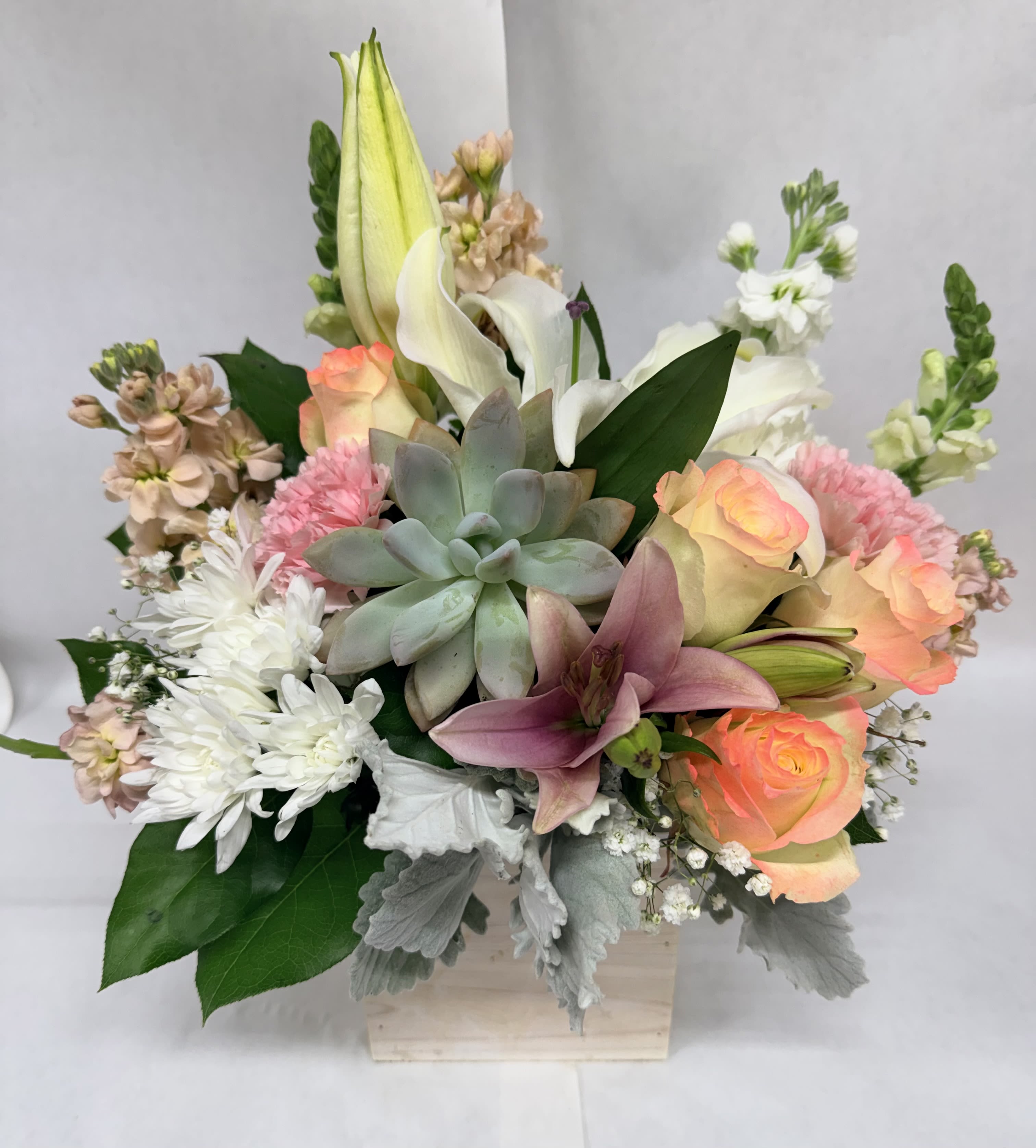 Desert Sunrise box wood Bouquet  - Pink and blush roses meet crisp white lilies in a soft, romantic Desert Sunrise box wood bouquet designed to make the person you're thinking of feel gently celebrated. This flower arrangement for birthdays, anniversaries, or sympathy brings a calm, uplifting presence-perfect when you want to send warmth without overwhelming fanfare.  The compact, layered design pairs white chrysanthemums and fragrant stock with a central succulent and broad green foliage for texture and contrast. Soft pastels of peach, blush, pink and white are arranged low in a light wooden box, giving the display a clean, rustic look that sits beautifully on a desk, mantel, or bedside table.   Thoughtfully composed to feel fresh and elegant, this box bouquet suits anyone who appreciates understated beauty-new moms, close friends, or someone needing comfort. The arrangement arrives ready to display; keep flowers in a cool spot away from direct sun and replenish water as needed to extend their life.