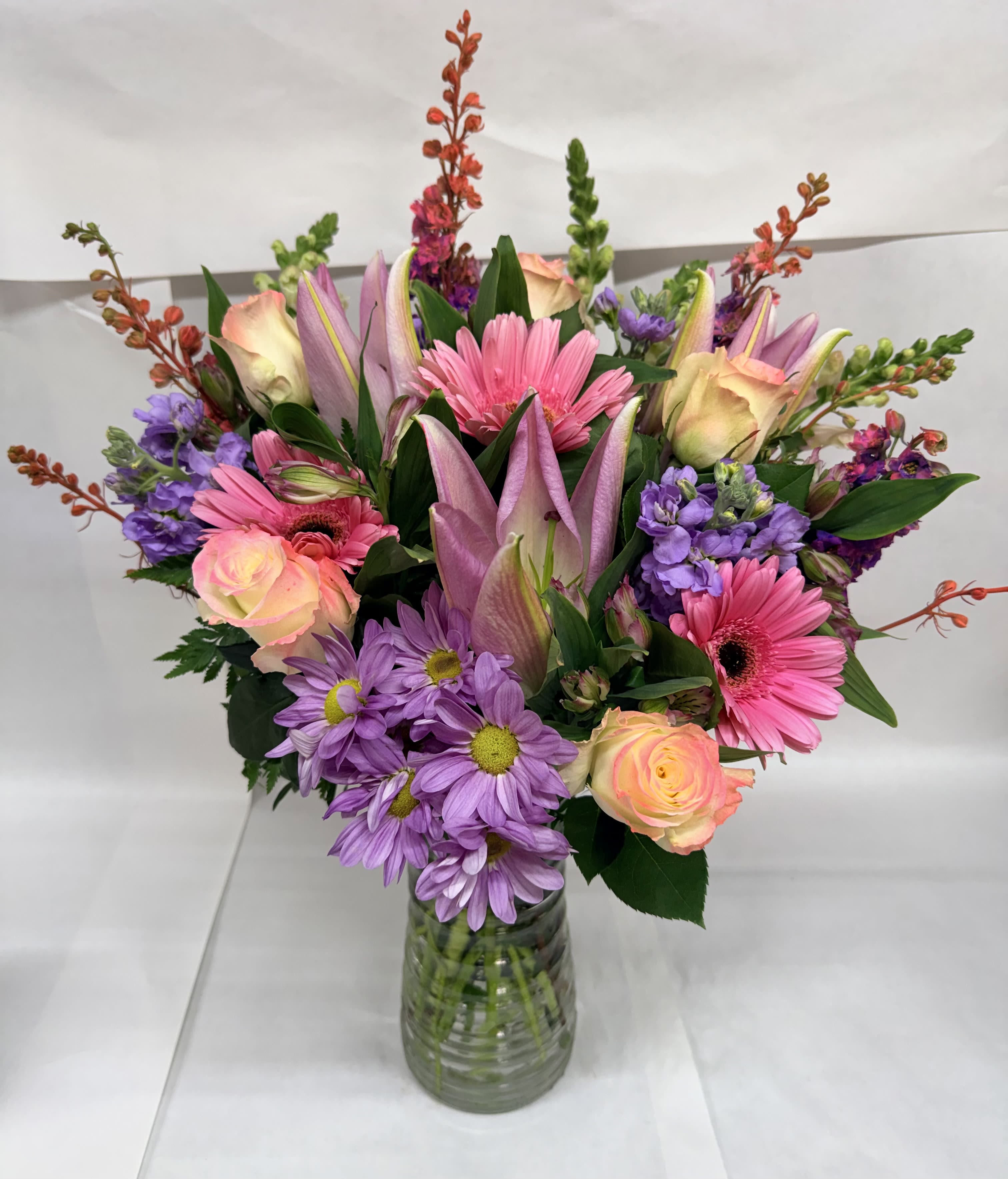 Always in my heart Bouquet - Soft pink gerbera daisies and pink roses take center stage in this Always in my heart bouquet, a cheerful flower arrangement perfect for birthdays, spring celebrations, or to tell someone you're thinking of them. Lavender daisy-like blooms and purple accents lift the palette into soft purples and creams, creating a warm, uplifting presence that will make the recipient feel truly appreciated.  This rounded design is arranged in a clear glass vase so the stems show through, keeping the presentation fresh and casual while letting the blooms speak for themselves. Tall accent spikes rise above the main cluster for height and movement, and the mix of gerbera daisies, roses, and complementary flowers gives a lively, layered texture that reads both joyful and thoughtful.  Ideal for a friend, mom, or colleague who deserves a bright moment, this bouquet sends a heartfelt message without being overdone-the sort of gift that brings a smile when it arrives. Vase included; trim stems and change water regularly to keep blooms looking their best.