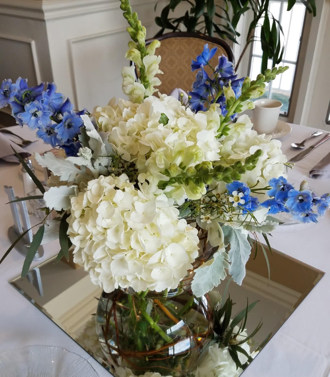 Centerpiece of white hydrangeas, snapdragons, and blue delphinium in a clear vase on a mirror.