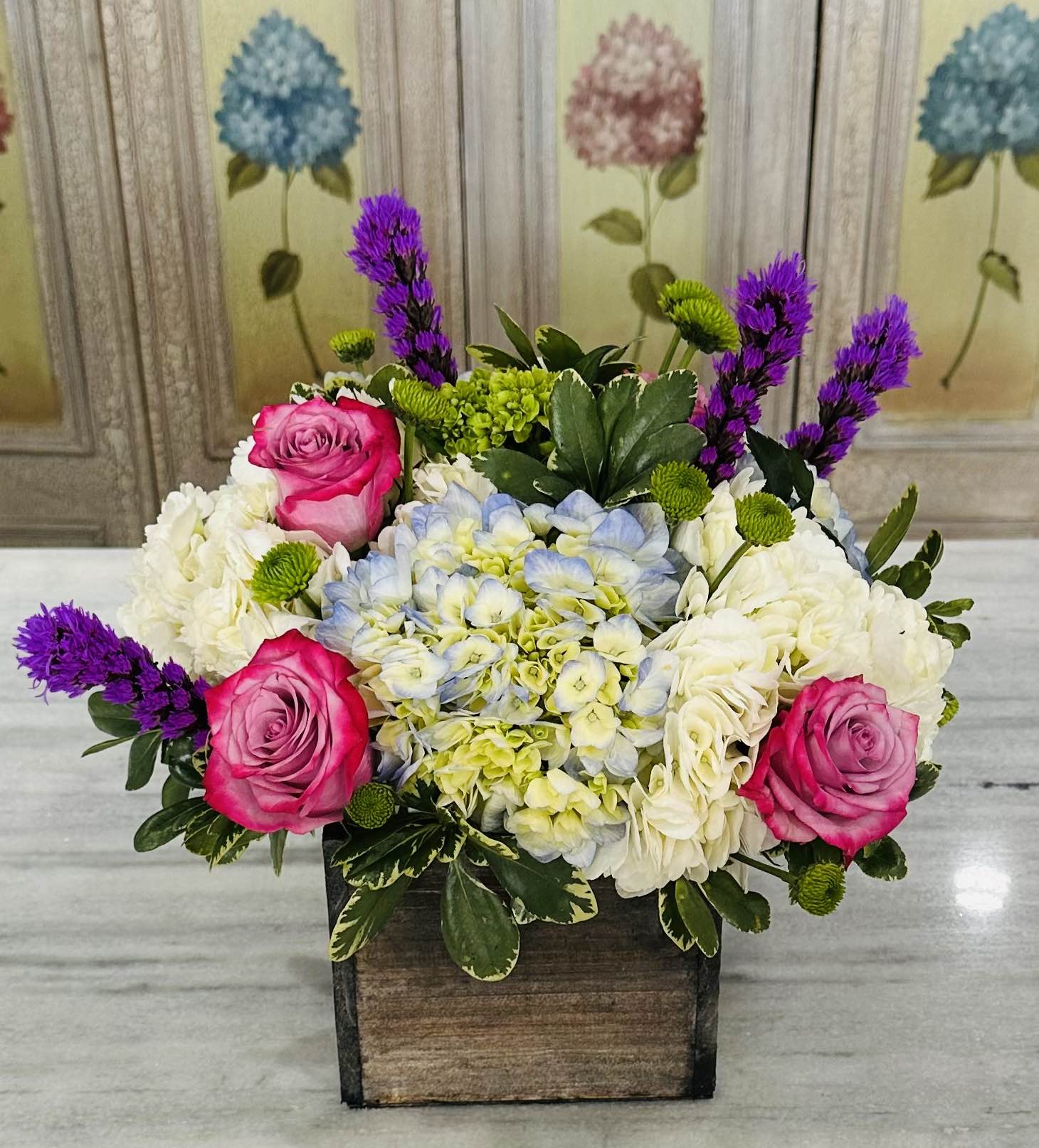 Low wooden box arrangement with blue hydrangeas, white blooms, pink roses, and purple accent flowers.