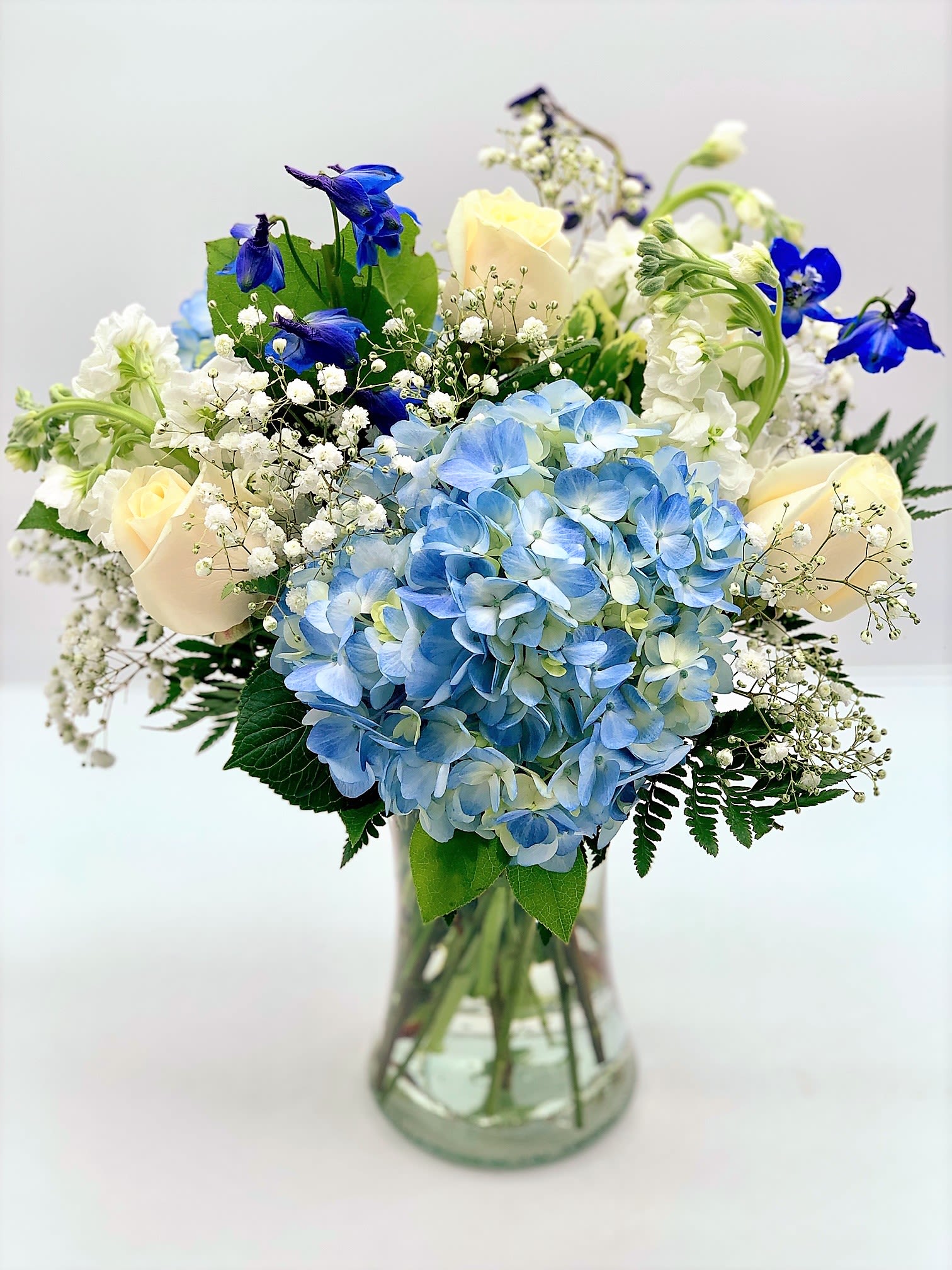 Vase arrangement with blue hydrangeas, cream roses, white blooms, and bright blue flowers