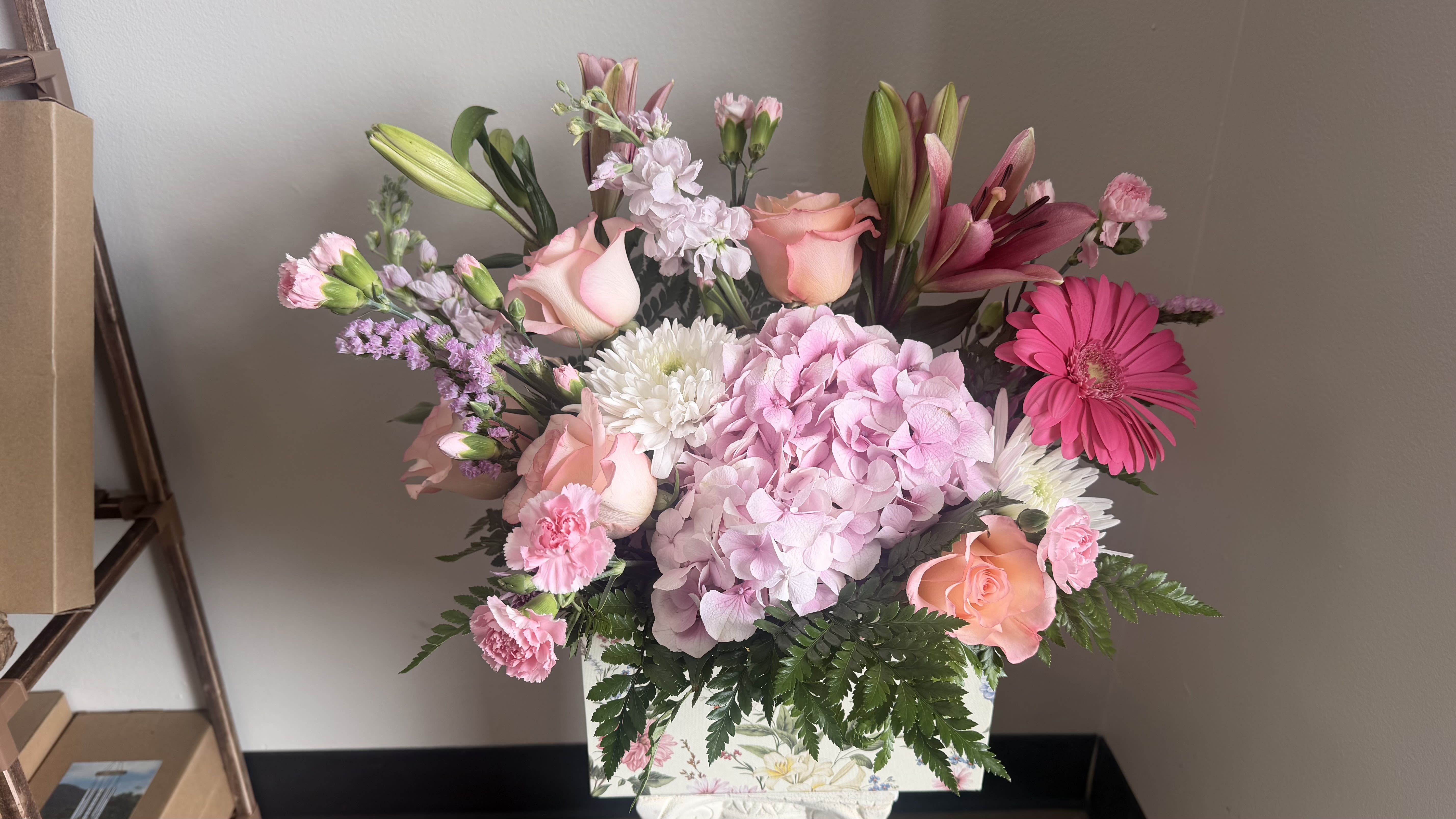 “Blush Garden Charm” - A lush cluster of pale pink hydrangea and blush roses gives this flower arrangement a soft, garden-fresh presence that will make her feel truly seen. Blush Garden Charm is a floral arrangement perfect for Mother's Day, birthdays, anniversaries, or a "thinking of you" surprise-the taller lilies and stock create an airy, uplifting silhouette while bright pink gerbera and white daisy-like blooms add cheerful pops of color for a joyful, feminine statement.  This mixed bouquet blends hydrangeas, roses, lilies, carnations, daisies, stock, and alstroemeria in a palette of pink, white, peach, and purple, presented in a decorative floral box for easy display. Textures range from the dense, cloudlike hydrangea to the long, elegant stems of lilies and stock, giving the piece pleasing height and depth. The arrangement reads as a generous, garden-style gift that suits spring and summer celebrations yet remains right for get-well wishes, housewarmings, graduations, sympathy, or holiday moments.  Choose this arrangement when you want the recipient to feel comforted, celebrated, or simply delighted-whether for Mother's Day, a wedding, Thanksgiving table accent, or "just because" kindness. The decorative box makes it simple to set out immediately; for best freshness, keep blooms in a cool spot and refresh the water if you transfer stems to a vase.