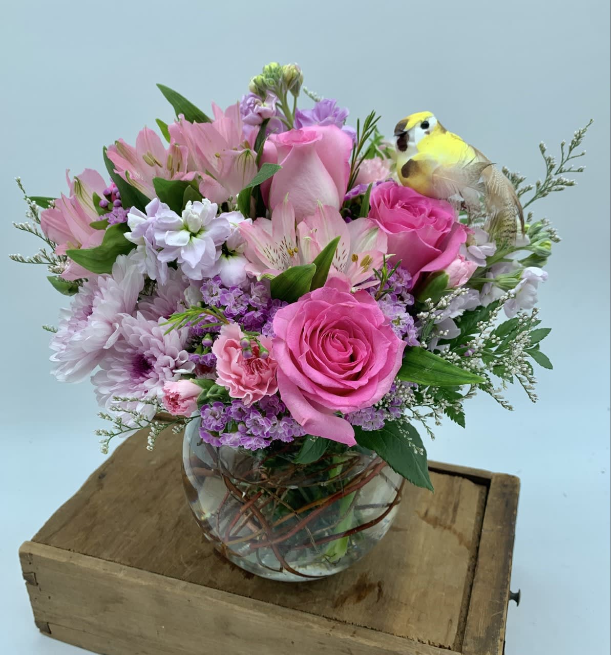 Round glass vase of pink and lavender flowers with a small yellow bird pick on a wooden box