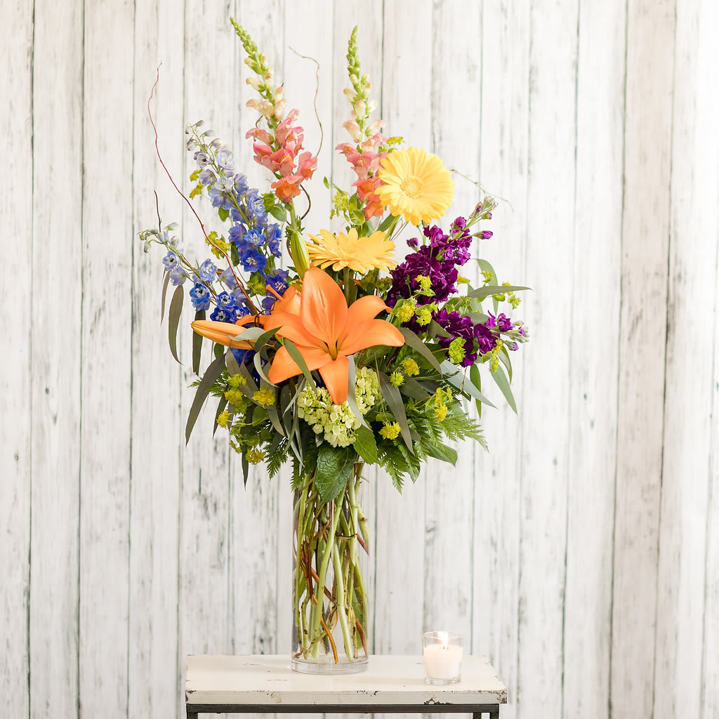 Tall mixed bouquet with orange lilies, yellow daisies, and purple blooms in a glass vase