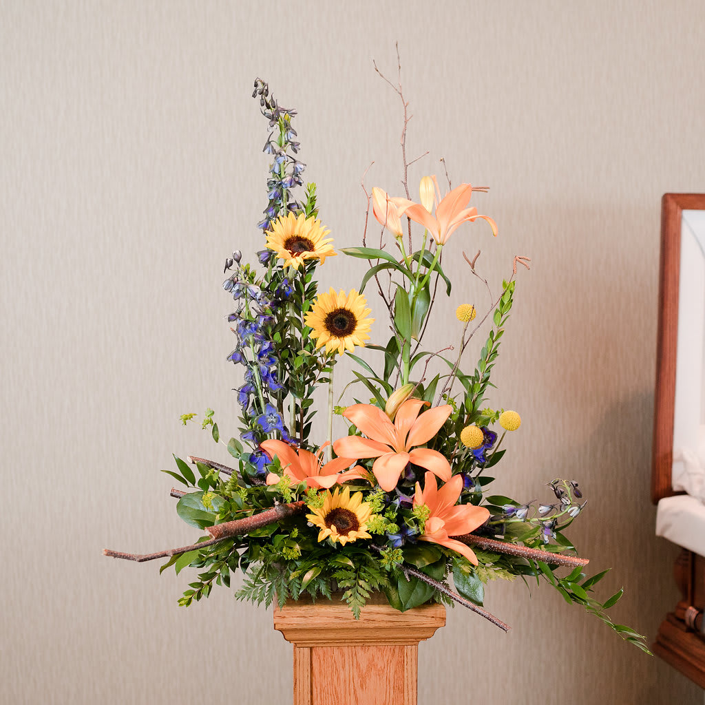Tall floral arrangement with peach lilies, sunflowers, and blue delphinium in a wooden pedestal vase