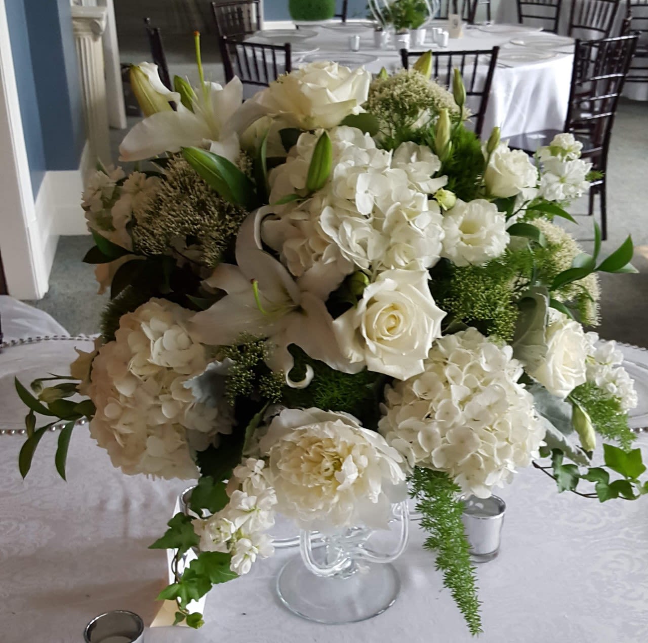 White floral centerpiece with roses, hydrangeas, and lilies in a clear vase