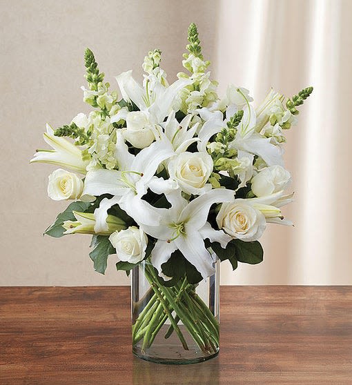 Arrangement of white lilies, roses, and snapdragons in a clear glass cylinder vase on a wooden table.
